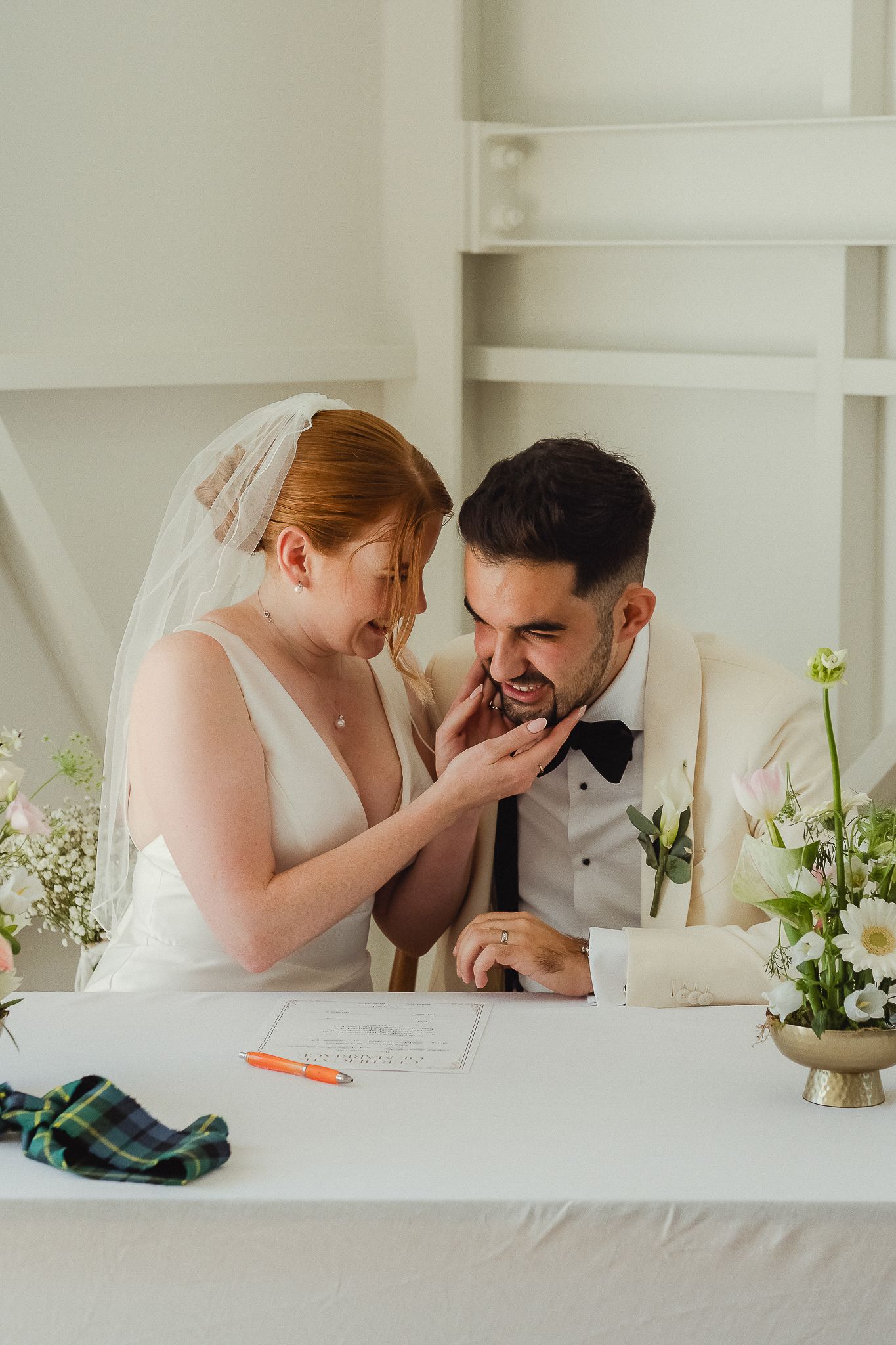 Bride and groom share joyful laughter while signing their marriage license at a white table decorated with flowers.