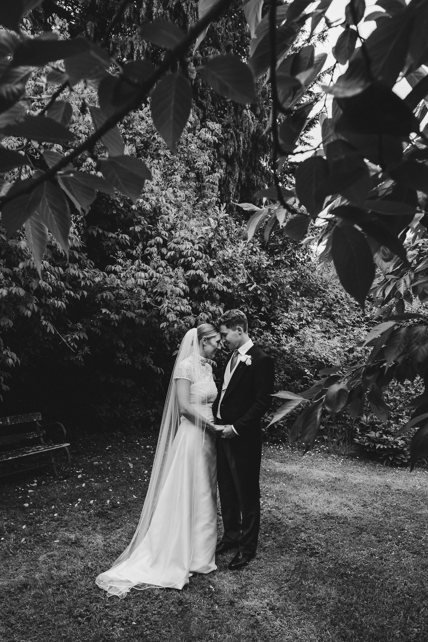 A black and white wedding photo of a couple embracing in a garden setting with lush foliage in the background.
