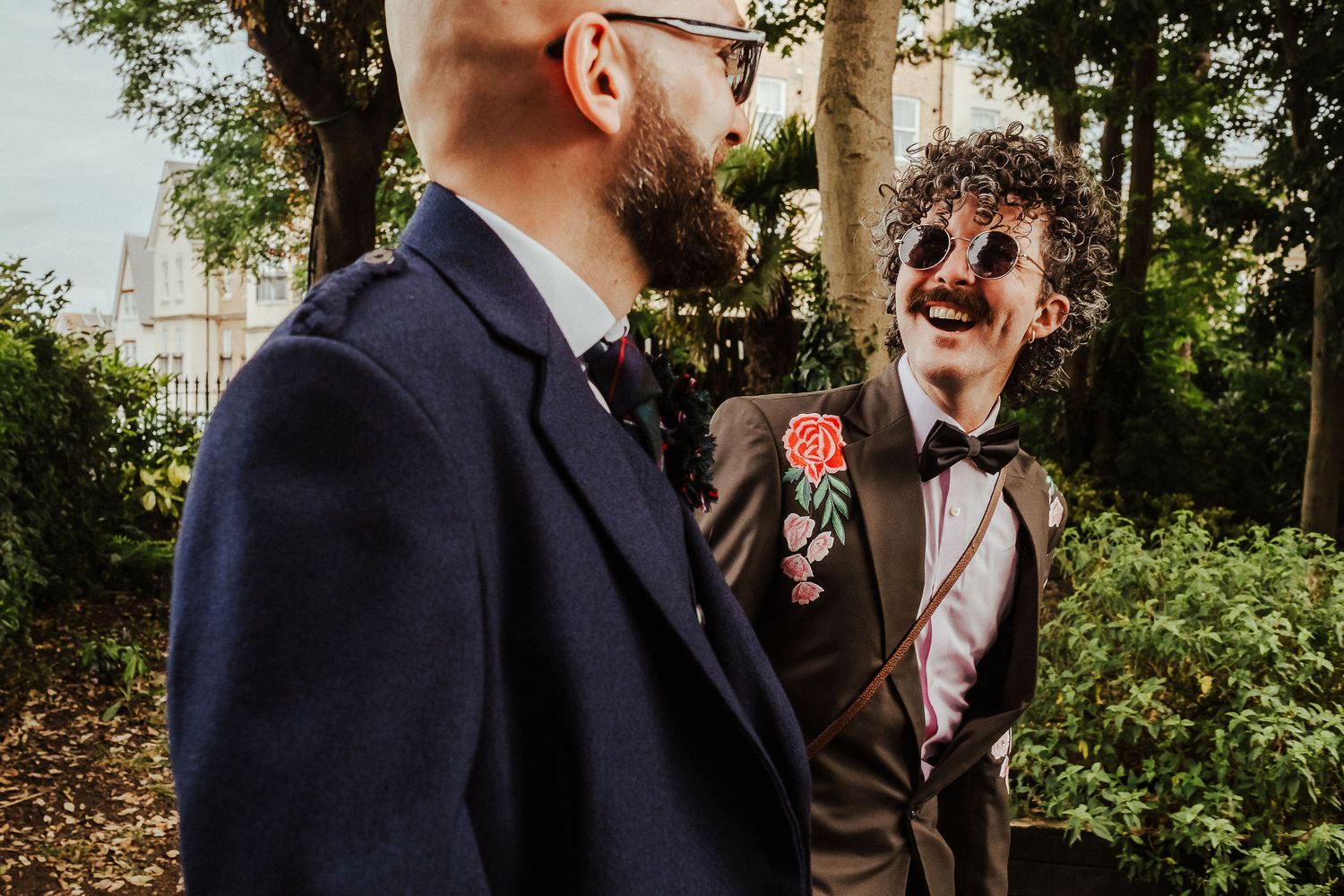 Two grooms in tailored jackets share a joyful moment outdoors, one laughing in round sunglasses with a floral boutonniere and bow tie.