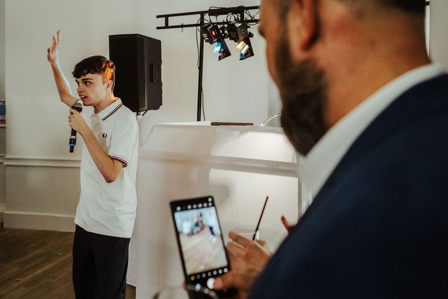 Young man in a white shirt and orange headphones holds a microphone and raises one arm while a bearded man films him on a smartphone.