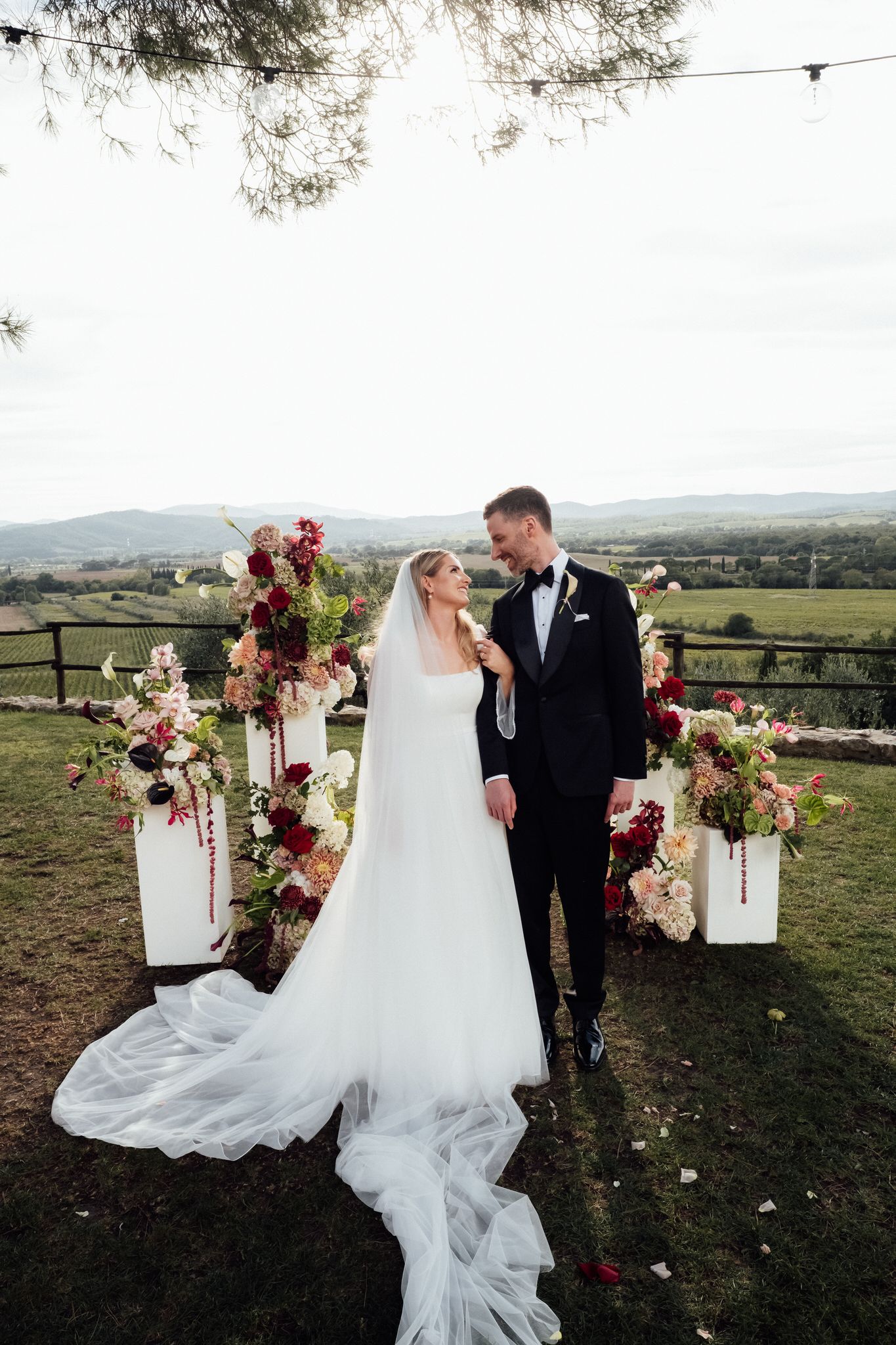 Bride in a long flowing veil and groom in a black tuxedo smile at each other beside floral arrangements at an outdoor countryside wedding.