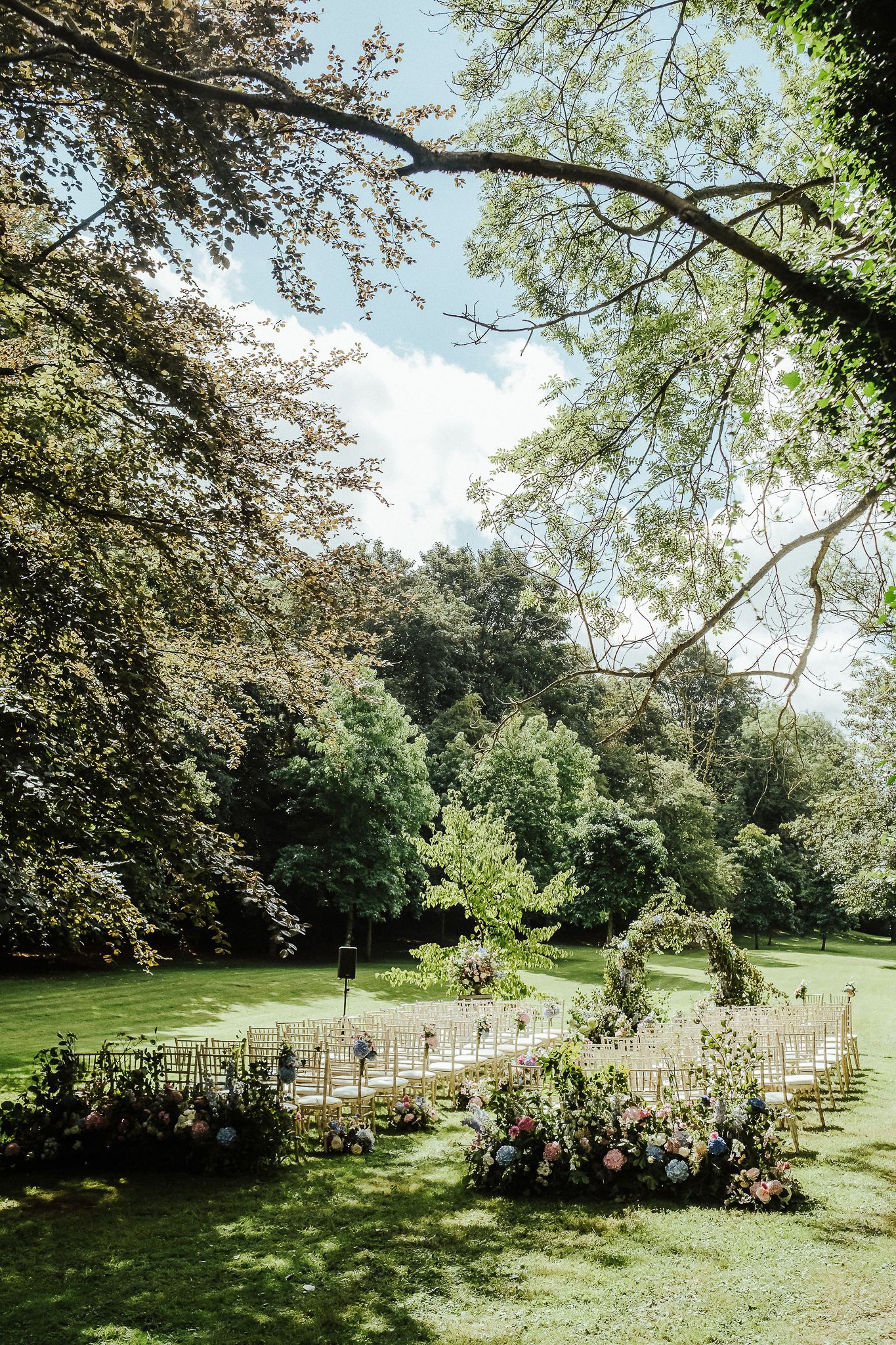 White chairs arranged in rows for an outdoor wedding ceremony in a lush garden setting beneath towering trees.
