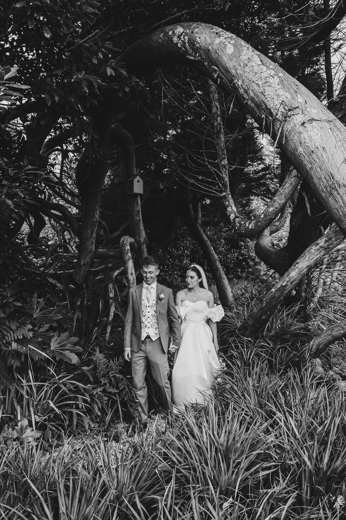Black and white wedding portrait of a couple walking through a lush garden path with overhanging trees and foliage.