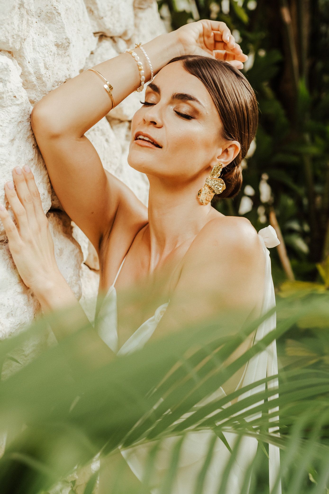 Artistic fashion portrait with tropical foliage in foreground and soft natural lighting creating a dreamy atmosphere.