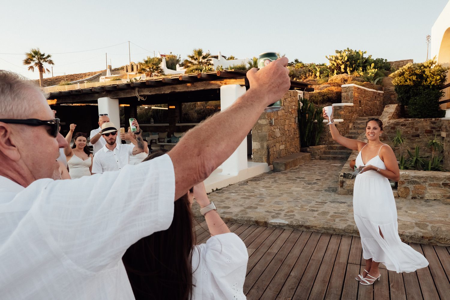 Guests in white attire celebrate outdoors, raising glasses in a festive toast at a sunny Mediterranean wedding.