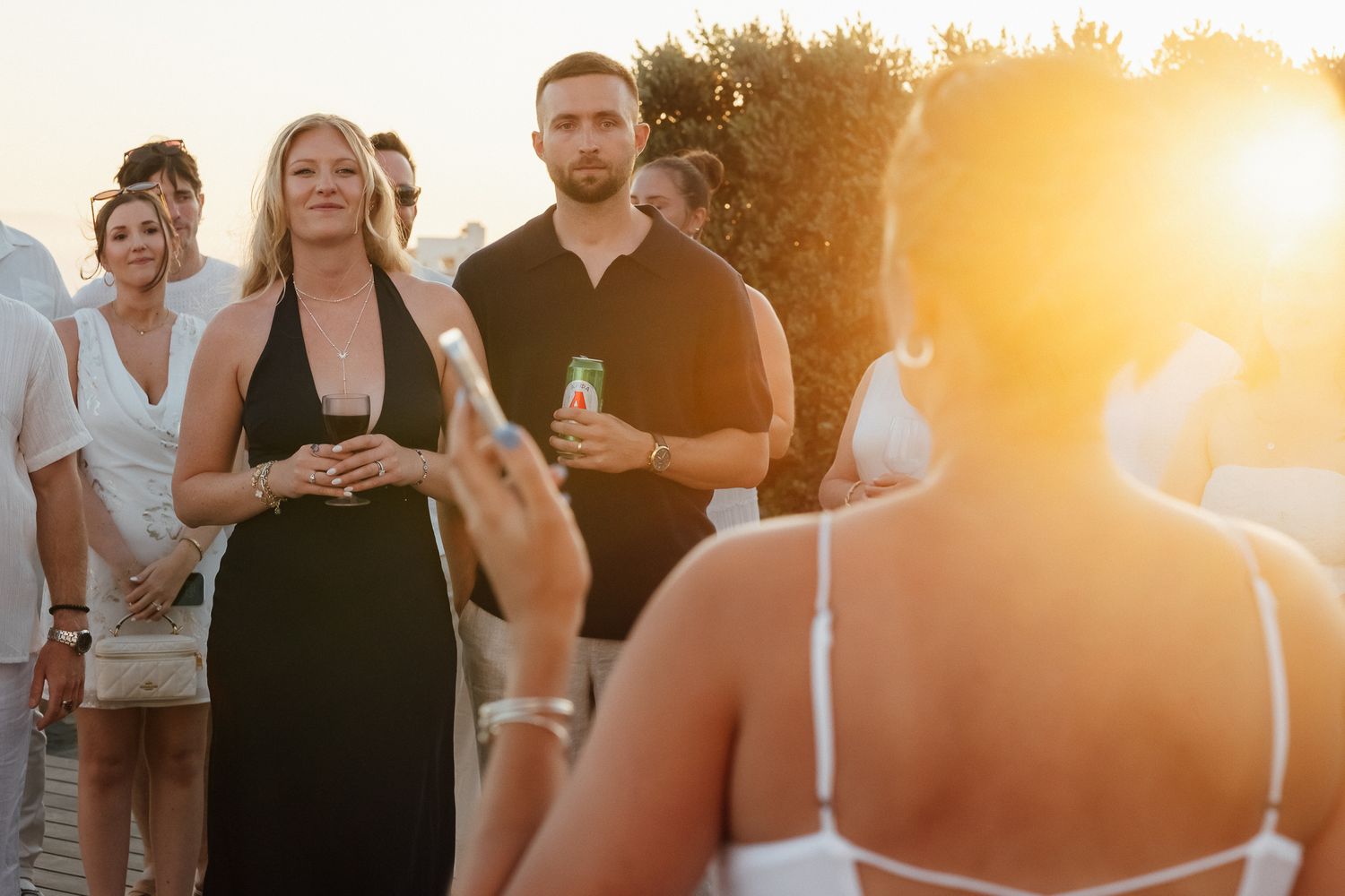 Guests socializing outdoors at a sunset gathering, woman in black dress holding a drink at a golden hour event.