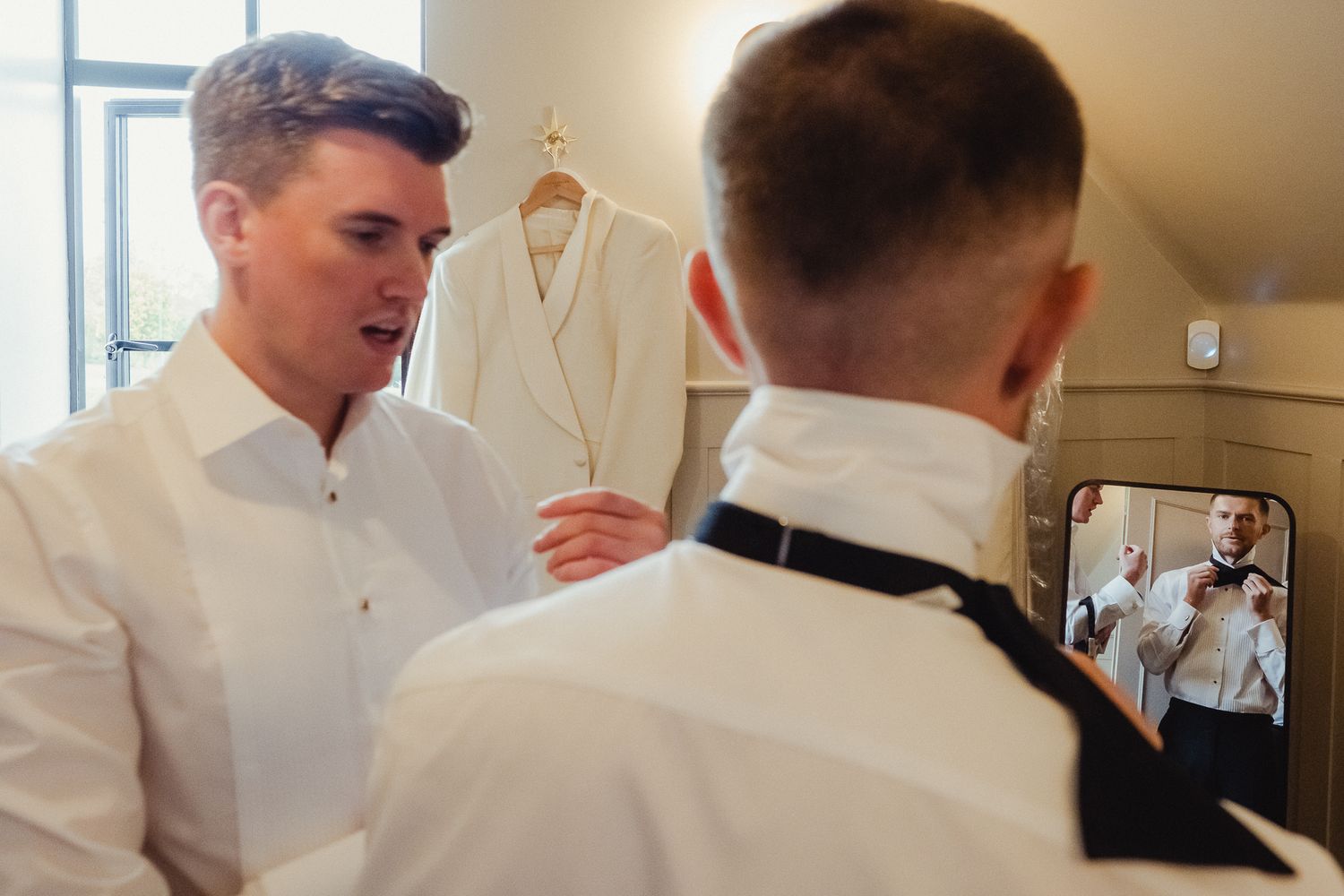 A person in a white dress shirt looks at a robe while someone takes a photo with a phone in the mirror.