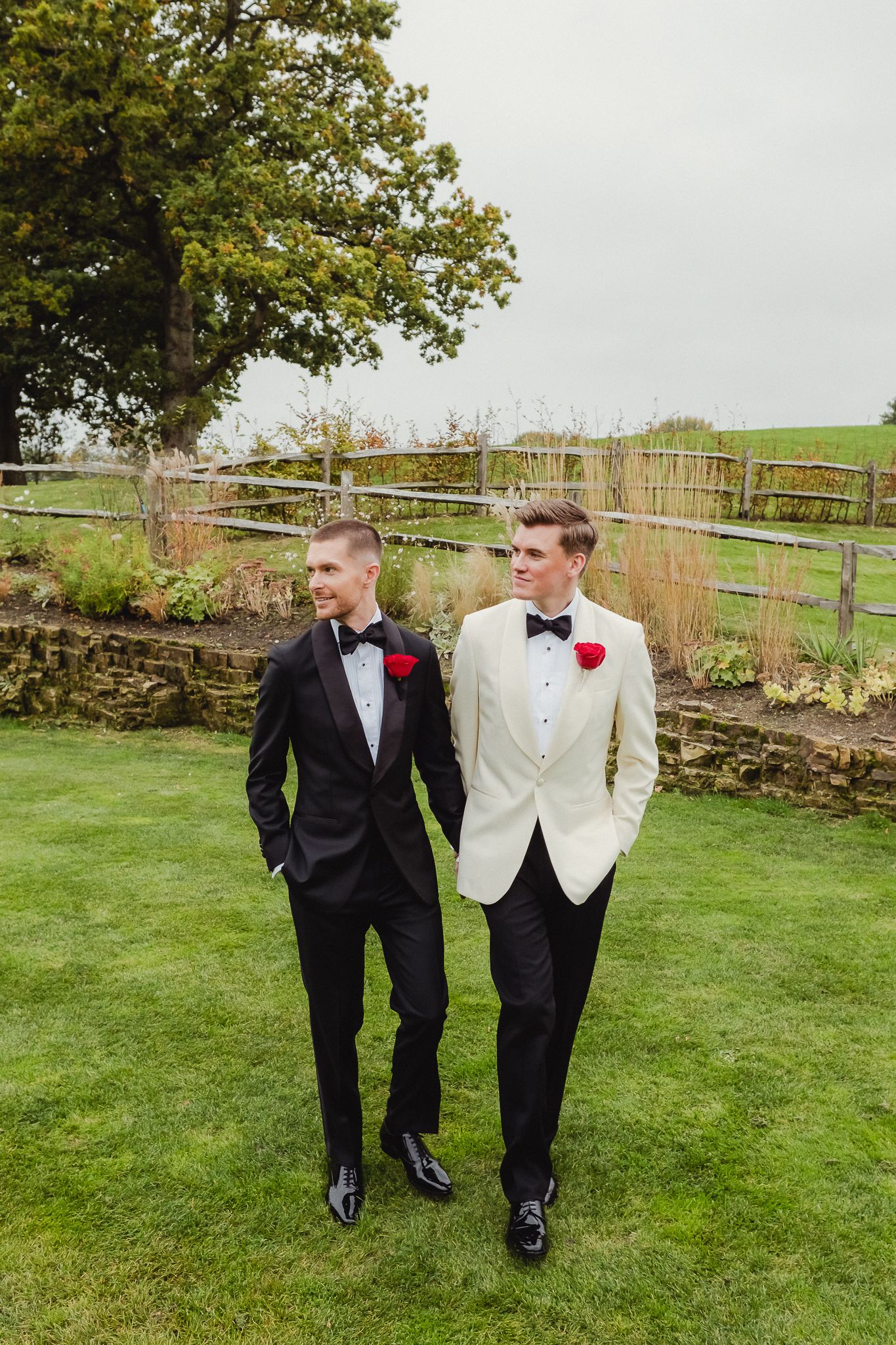 Two people in formal attire walking and laughing together on a green lawn with stone walls and autumn trees.