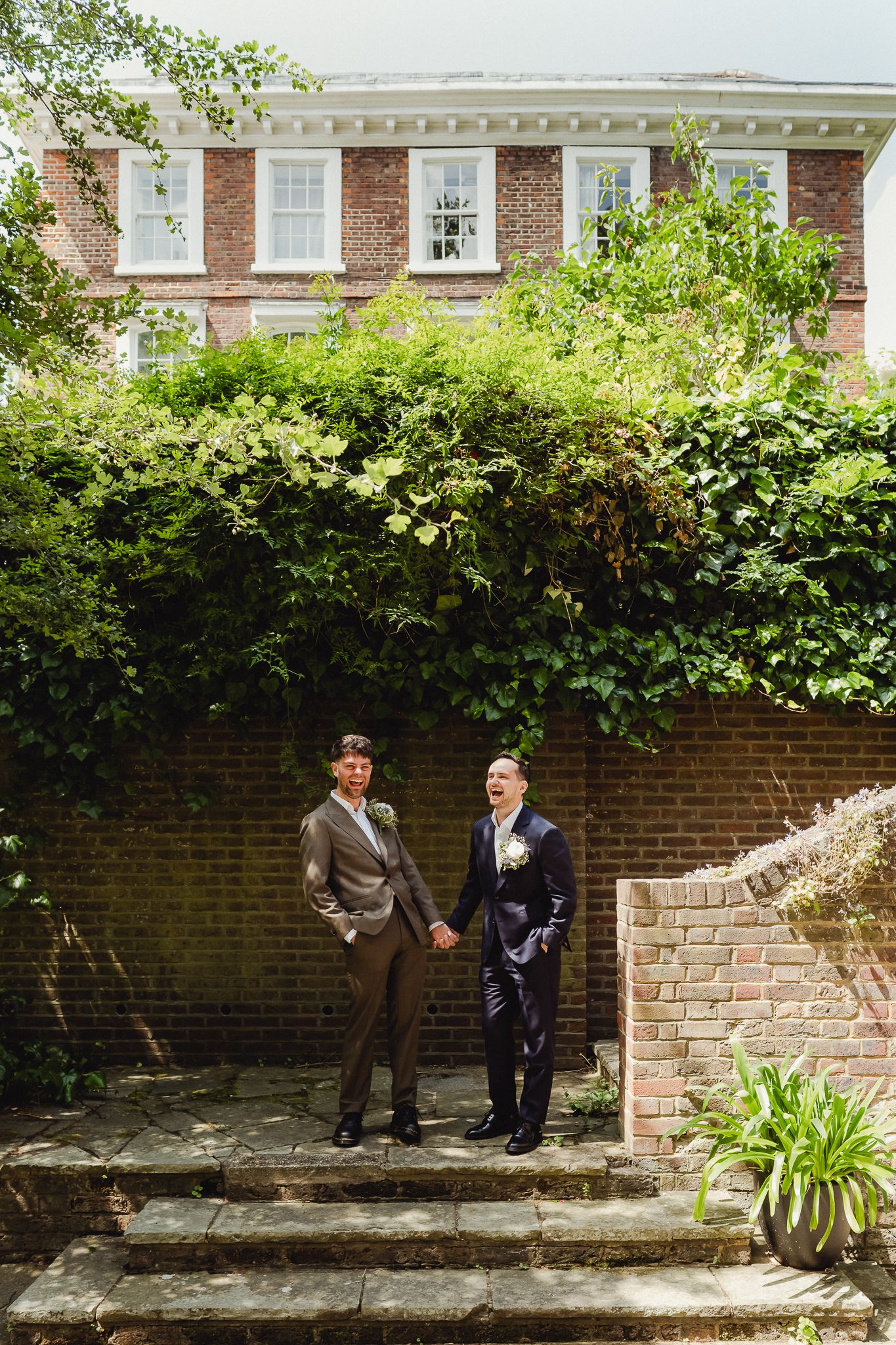 Two people in formal attire having a conversation while standing against a brick wall with greenery above.