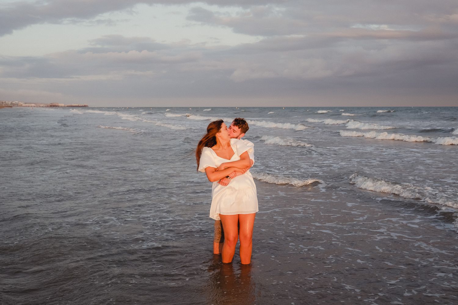 A series of romantic beach photos at sunset shows a couple in coral-colored outfits embracing by the ocean waves.