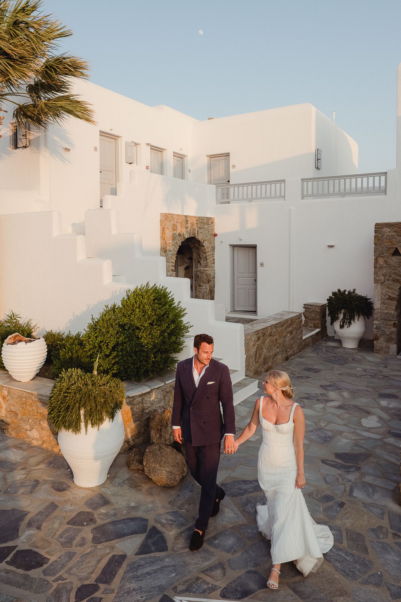 Couple walking along a Mediterranean-style stone path surrounded by white buildings and potted plants at sunset.