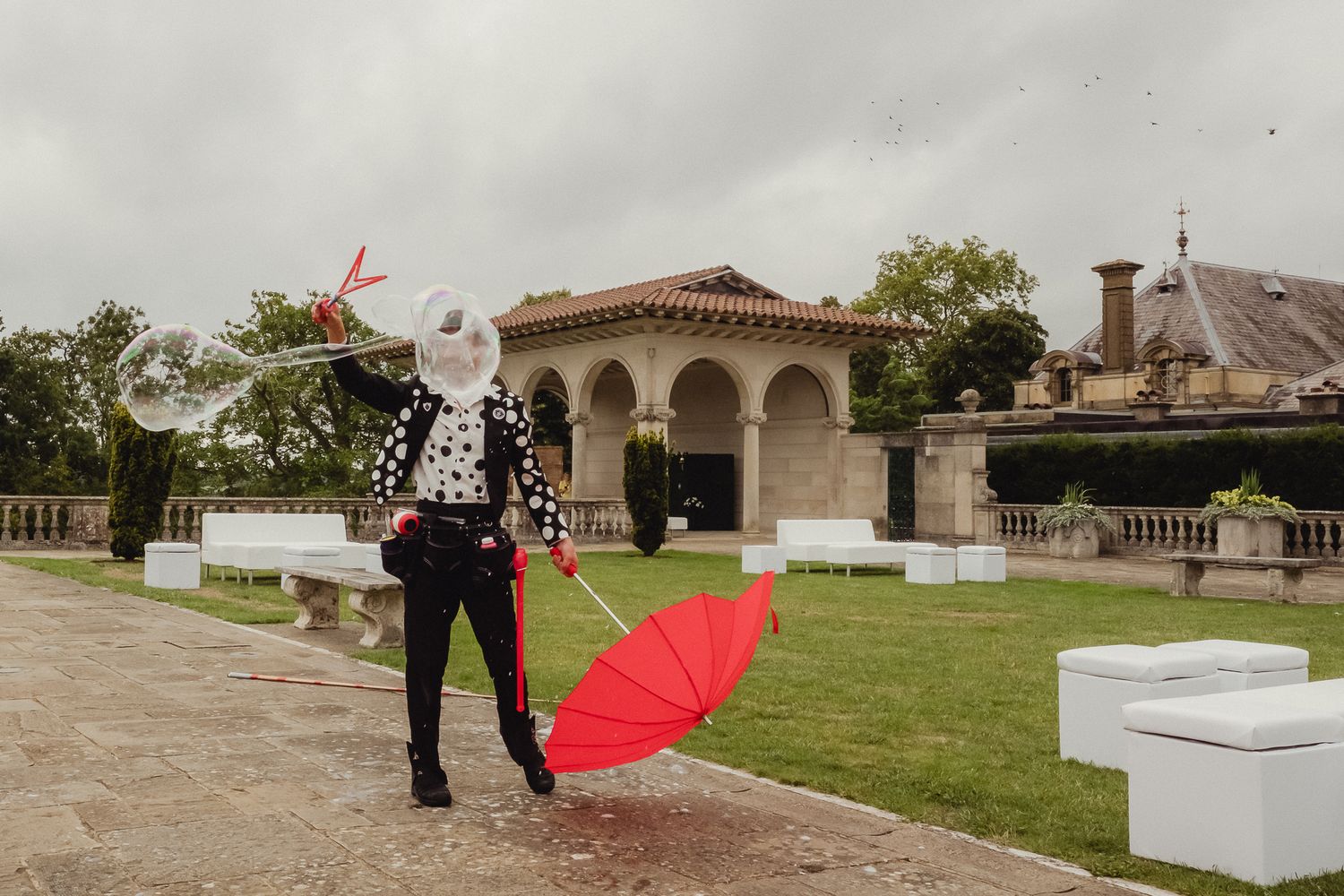 Person in black outfit poses with red umbrella in front of Mediterranean-style arched building on landscaped grounds.