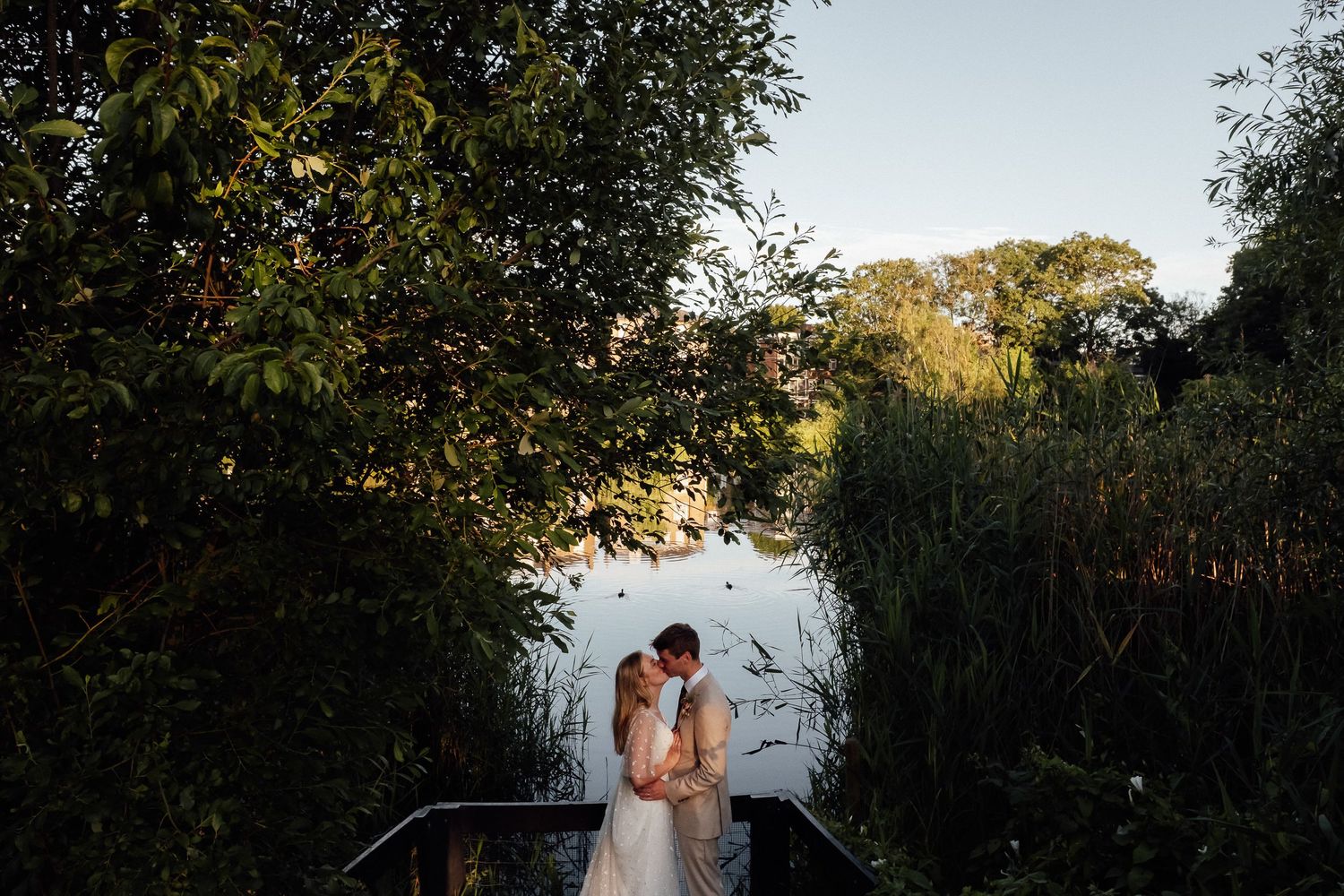 Bride and groom embrace on a wooden dock framed by lush green trees, overlooking a calm lake at golden hour.