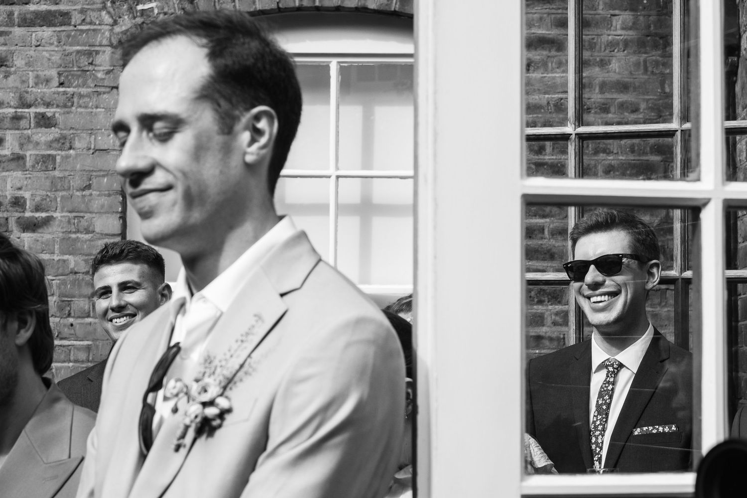 Black and white wedding photo of a smiling groom with a floral boutonniere as guests laugh, one wearing sunglasses reflected in a window.