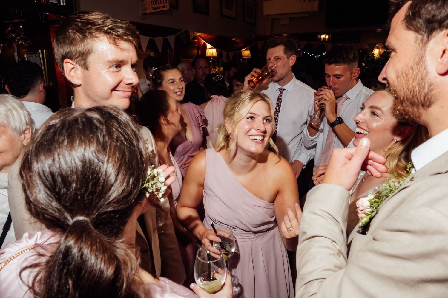 Smiling woman in a blush dress holds a wine glass while laughing wedding guests celebrate and clap around her at a lively reception.