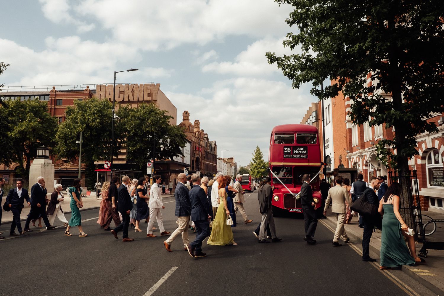 Wedding guests in formal attire crossing a sunny city street past a red double-decker bus, with trees and brick buildings lining the road.