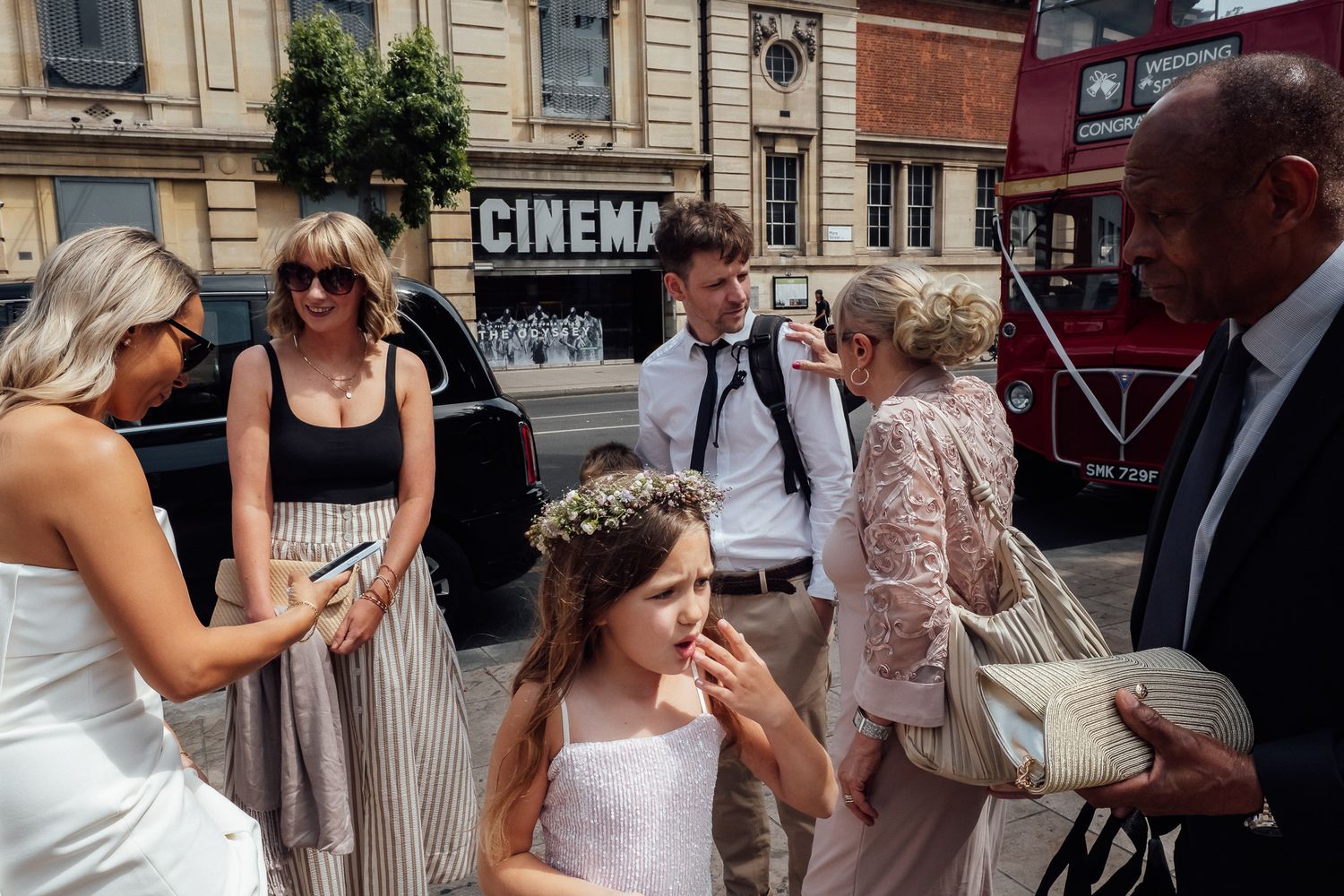 Young girl in a floral flower crown stands among wedding guests outside a cinema while a red double-decker bus passes in the background.