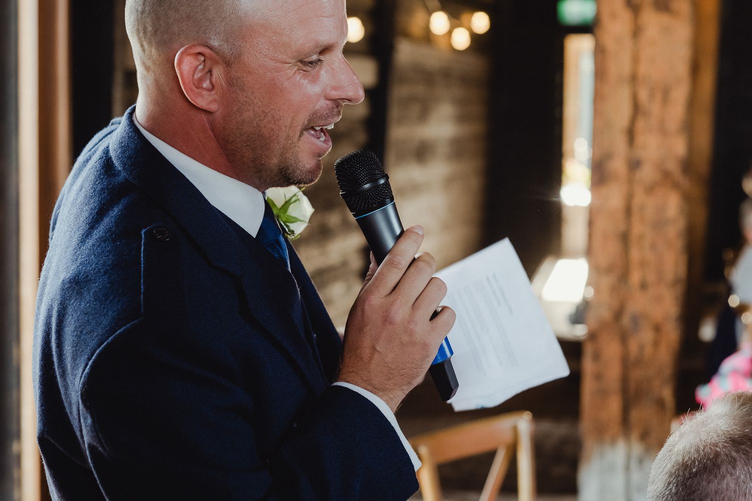 A person gives a heartfelt speech at a rustic wedding reception venue with wooden beams and warm lighting.