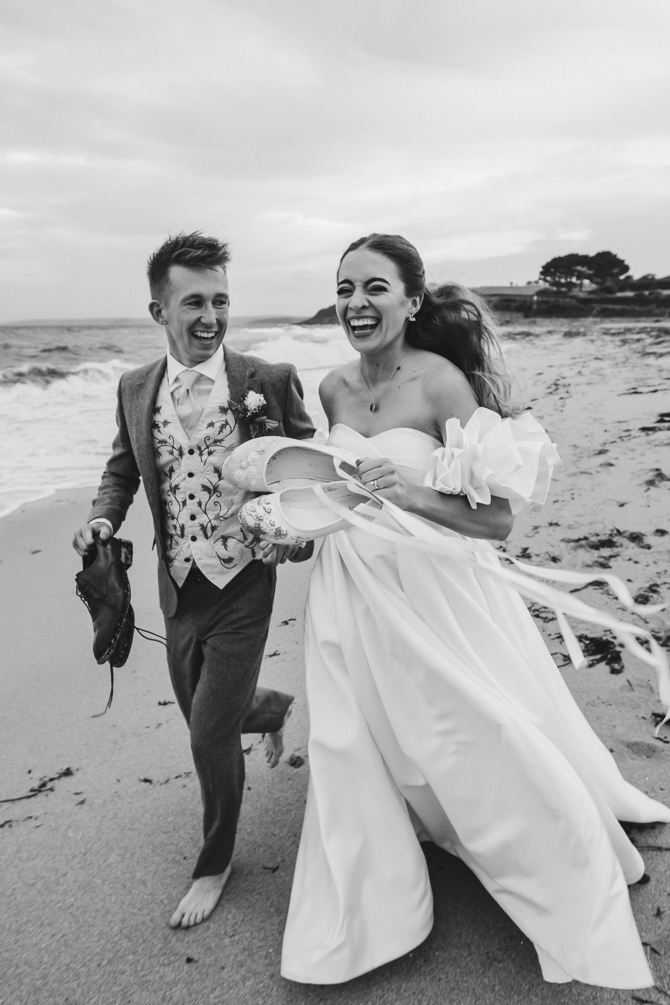 A joyful couple shares candid moments on a cloudy beach, with the bride's white dress flowing in the wind.