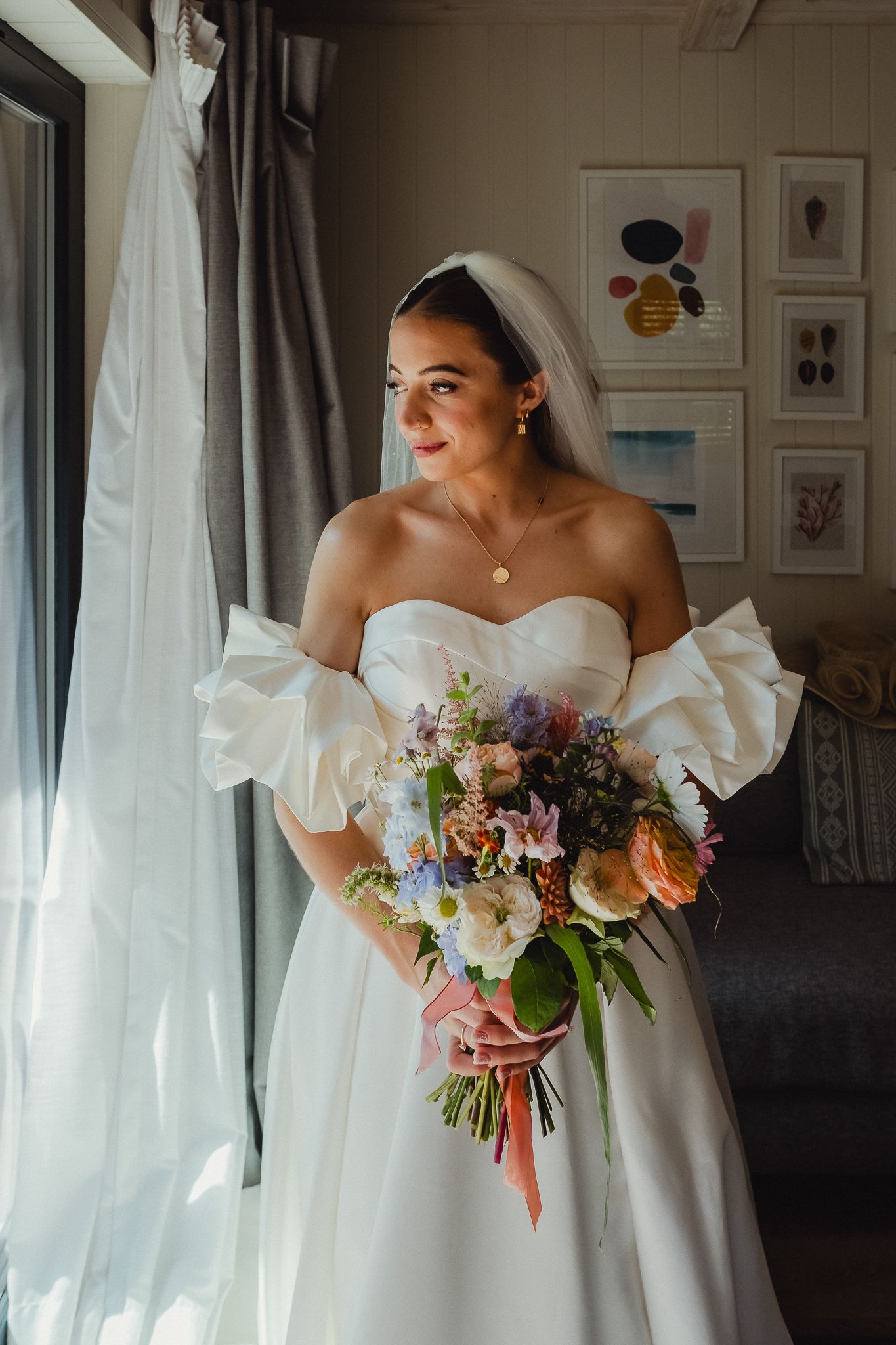 Bride in elegant white strapless wedding gown holds vibrant bouquet of mixed flowers near window with soft natural light.