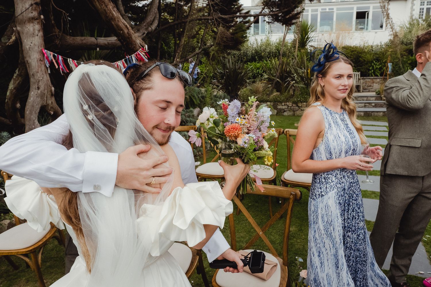 A joyful outdoor wedding celebration with guests dancing and embracing in a garden setting.