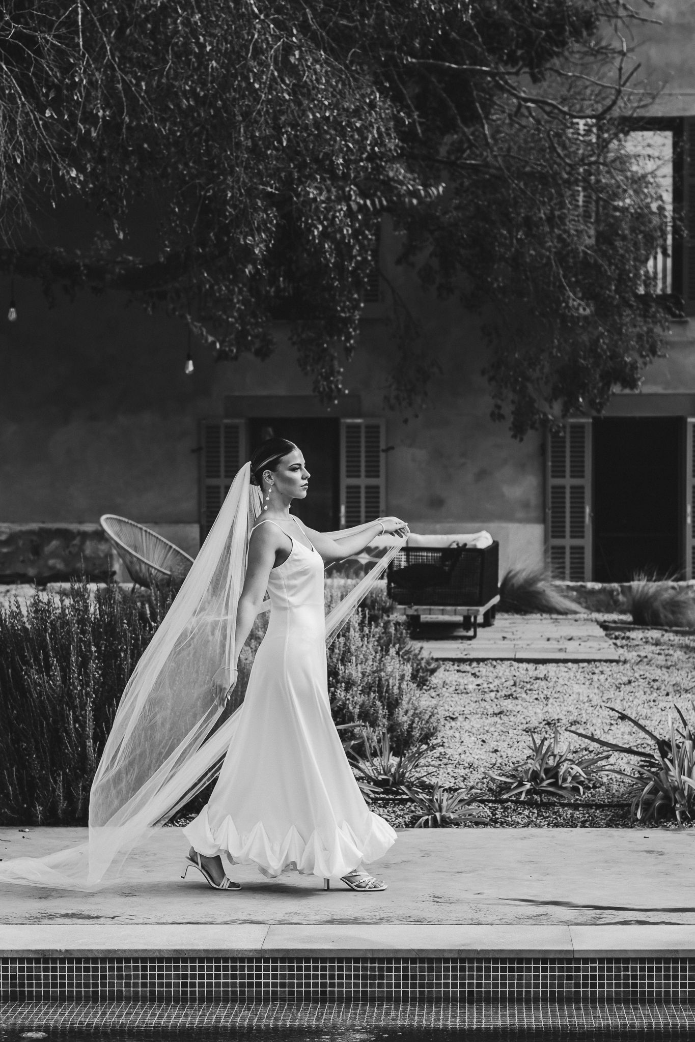 A bride in a flowing white gown and veil performs graceful dance movements by a pool in black and white photography.