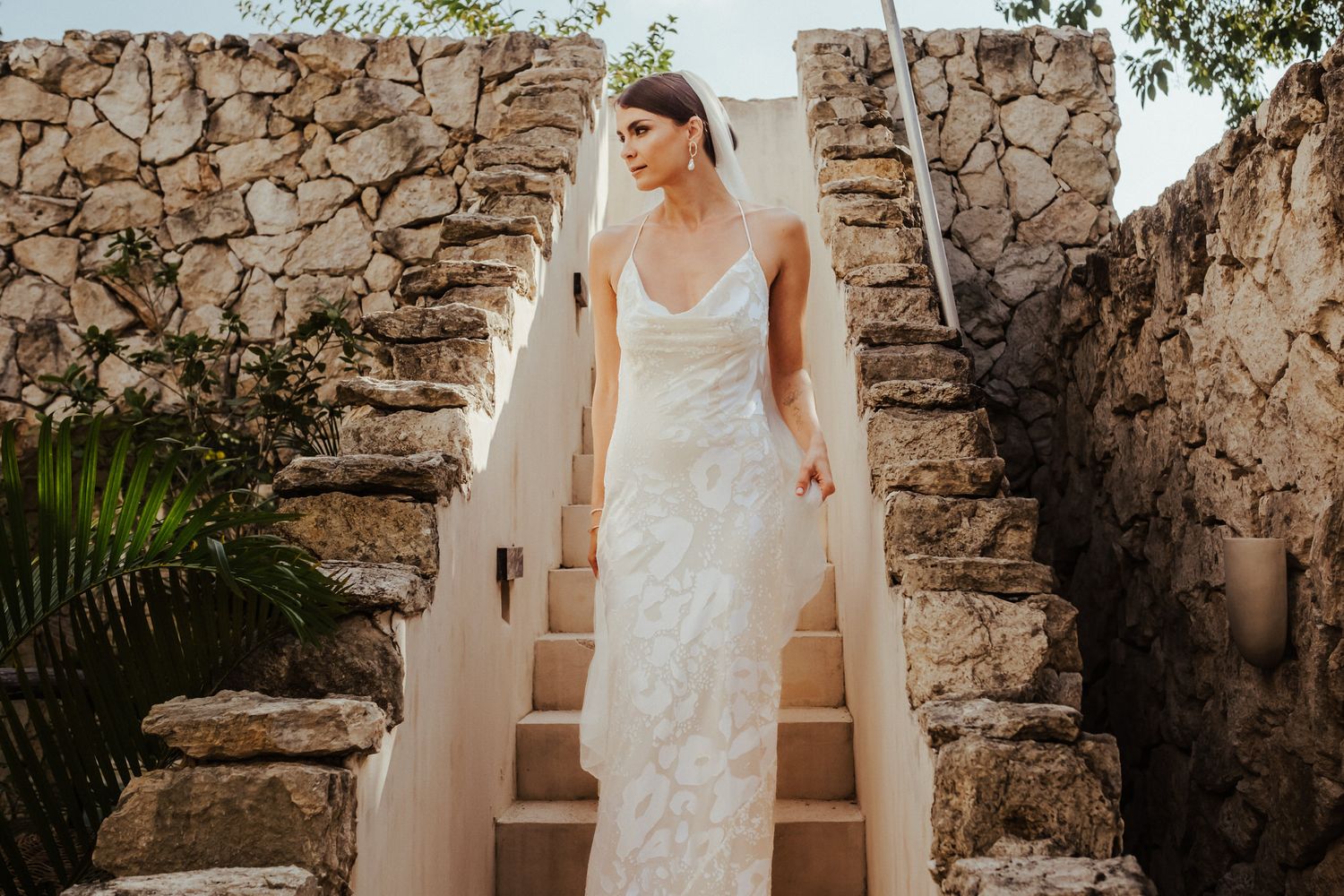 A person in a white lace wedding dress poses against a rustic stone wall background.