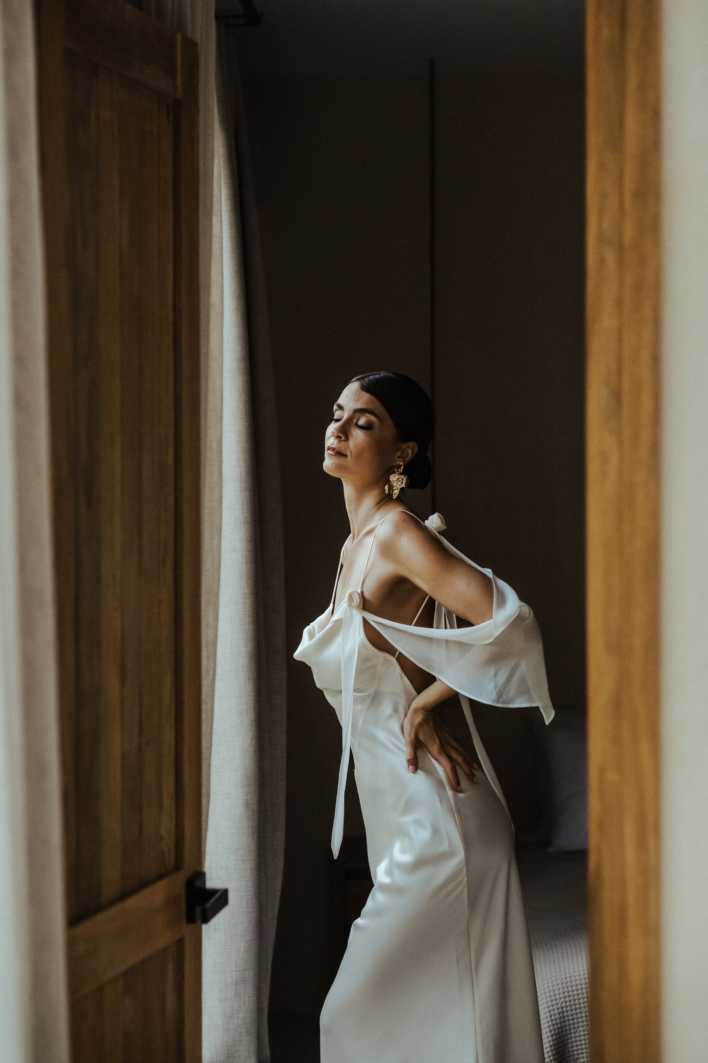 Woman in white dress poses elegantly between wooden columns in moody dramatic lighting.