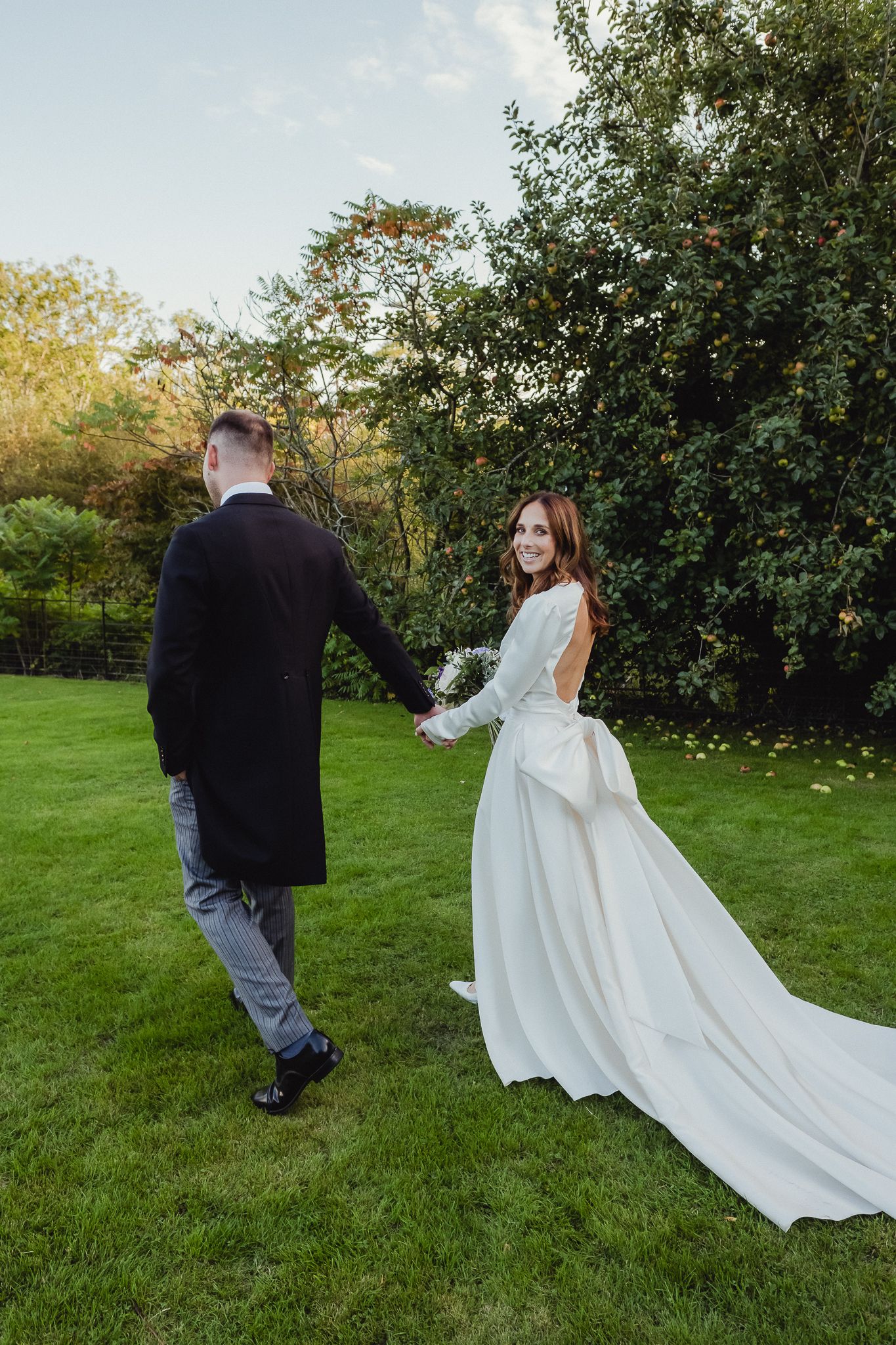 A couple in wedding attire walks together on a grassy lawn with the bride's white dress train flowing behind them.