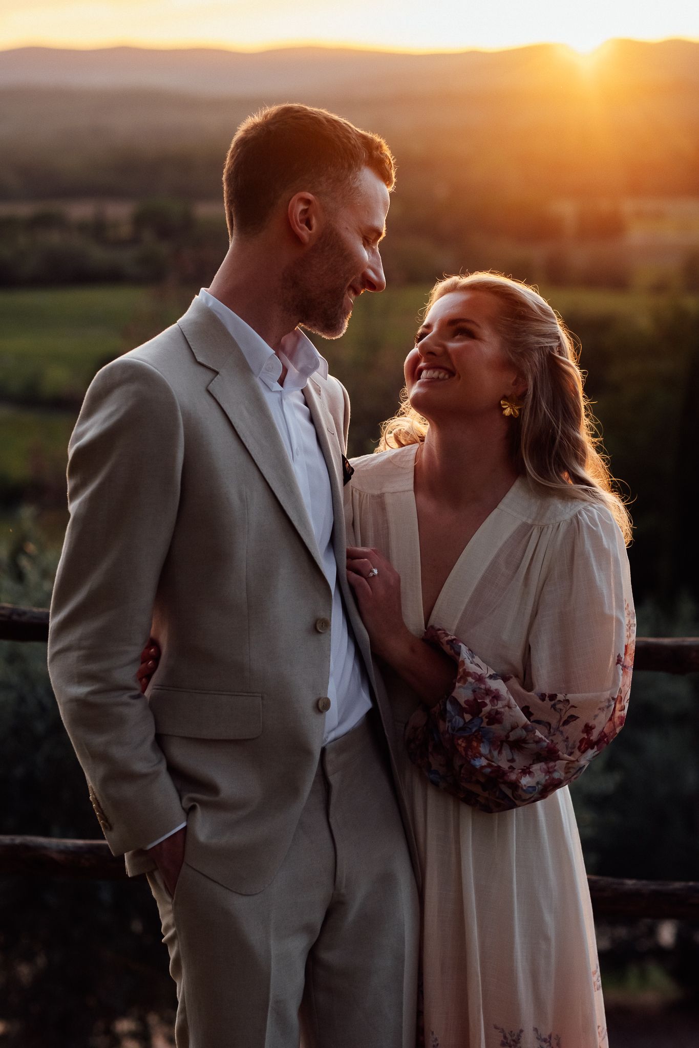 A smiling couple in light linen attire embraces at golden hour on a rustic overlook with vineyard hills and a glowing sunset behind them.