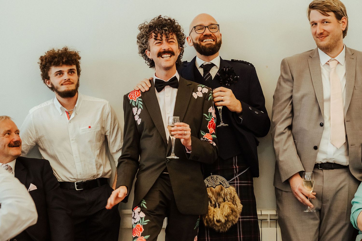 Smiling groomsmen in suits laugh together at a wedding, with a curly-haired man in a floral-embroidered jacket and bow tie at the center.