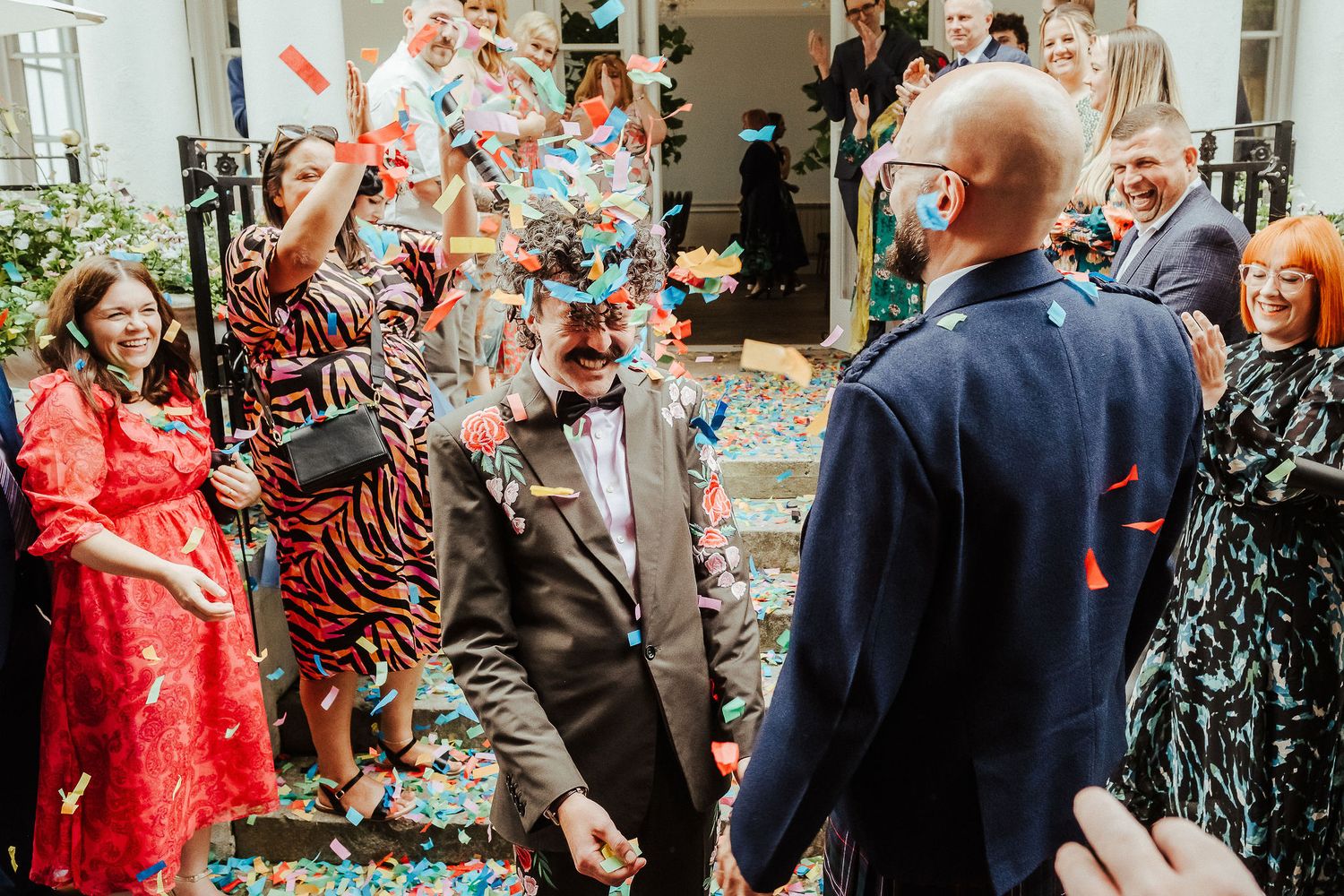 Wedding guests cheer and throw colorful confetti over a smiling groom in a brown suit with a bow tie outside a venue.