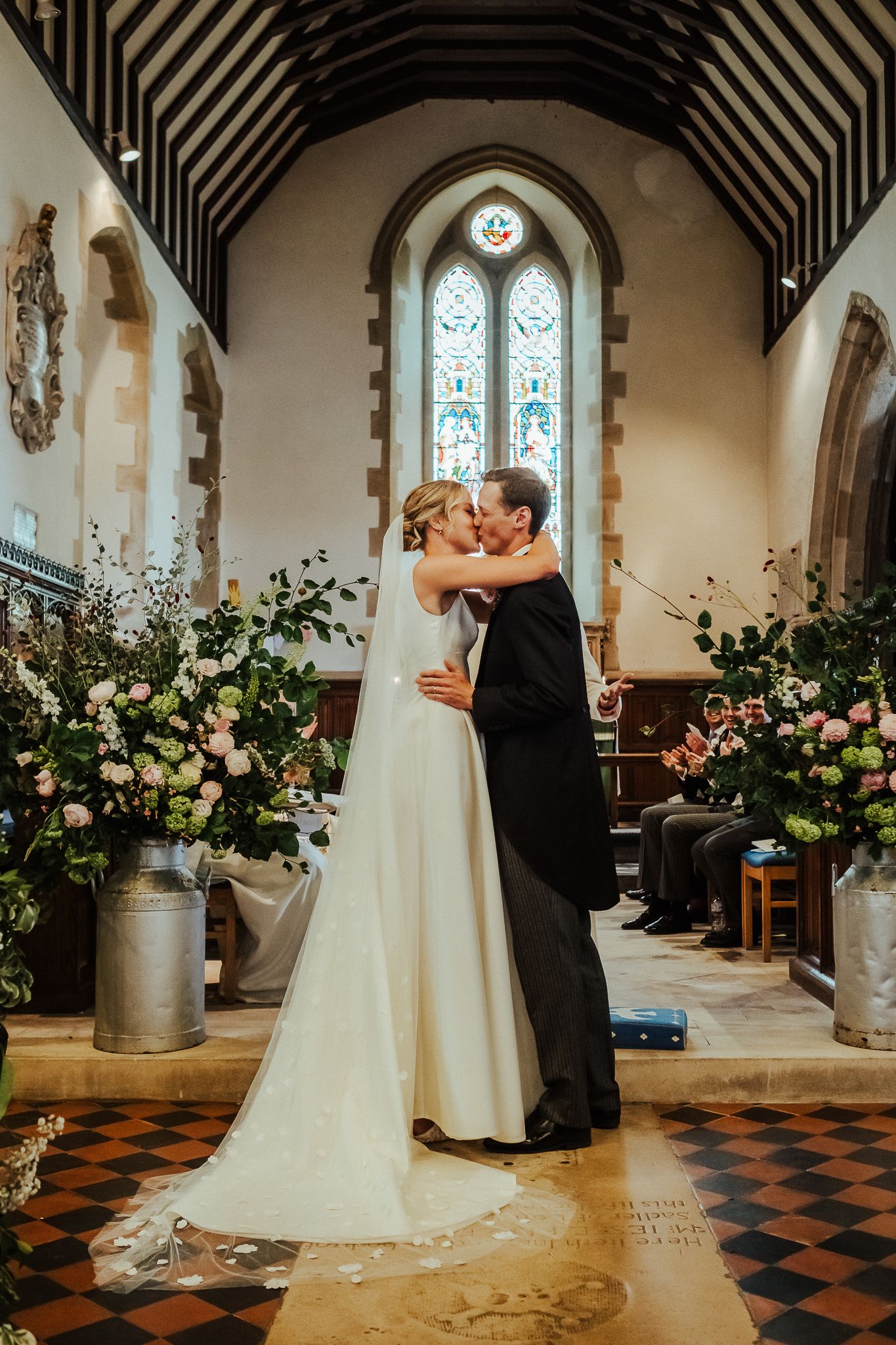 A romantic first kiss between newlyweds in a church with beautiful floral arrangements and stained glass windows.