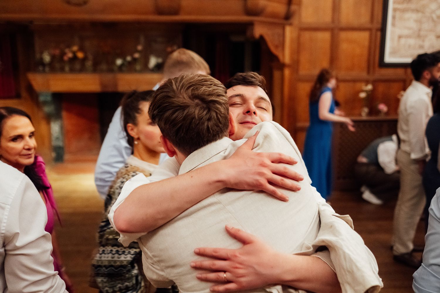 Two young men share a warm embrace at a wedding reception in an elegant wood-paneled venue.