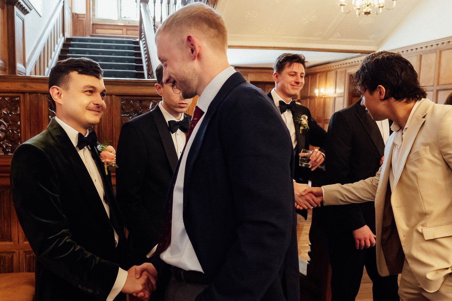 Men in formal tuxedos shaking hands and socializing at an elegant wedding reception in a wood-paneled venue.