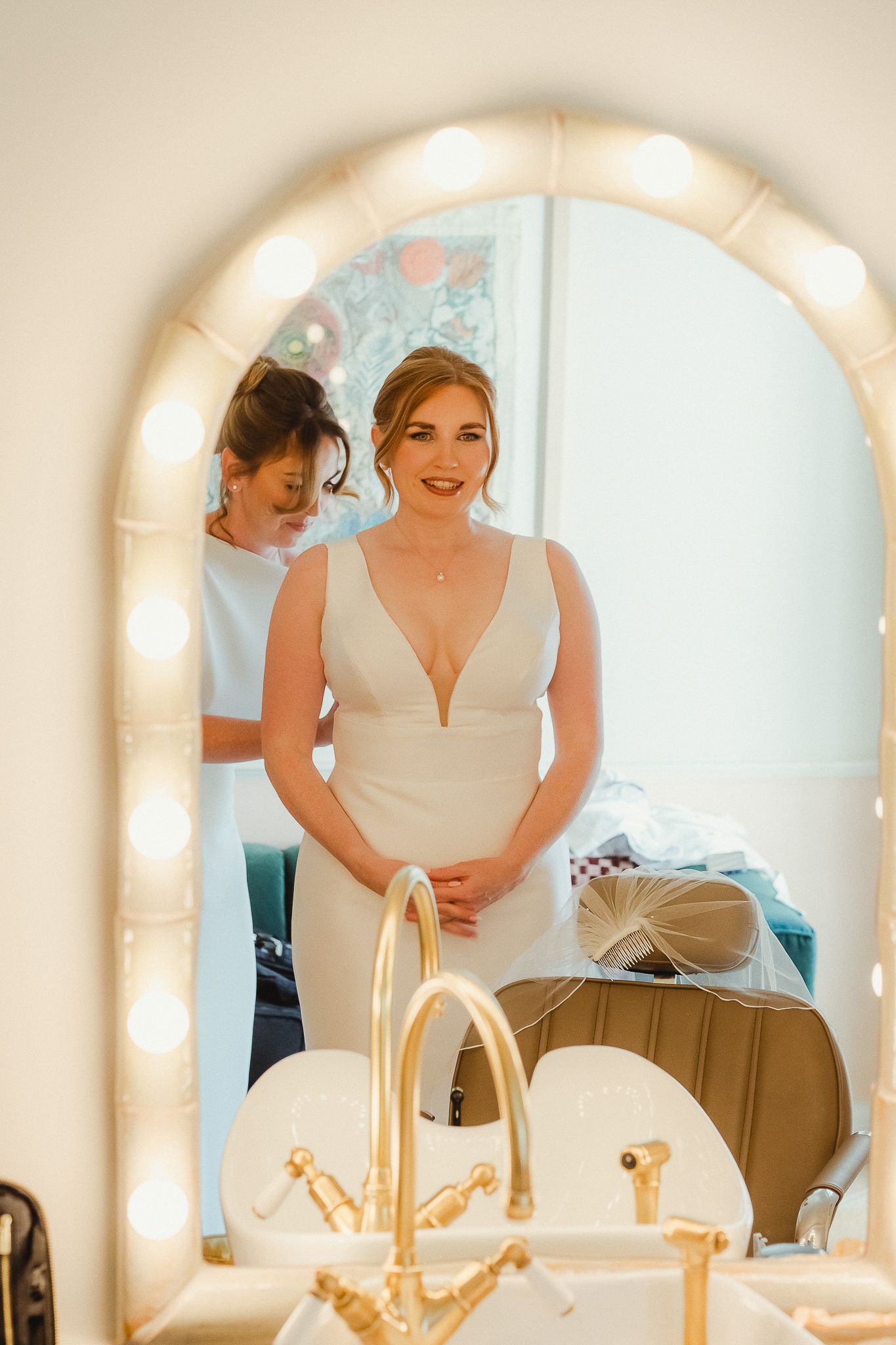 Reflection in a vanity mirror shows someone in a white wedding dress adjusting their gown in a dressing room.