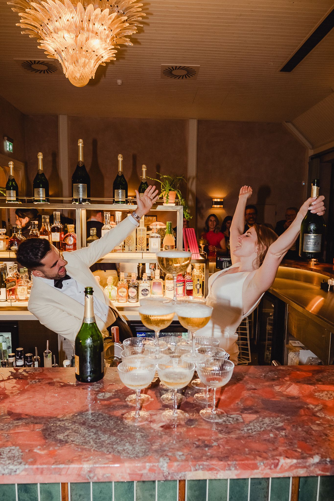 Sequence of cocktail glasses with splashing liquids and dramatic lighting on a bar counter with elegant chandeliers above.