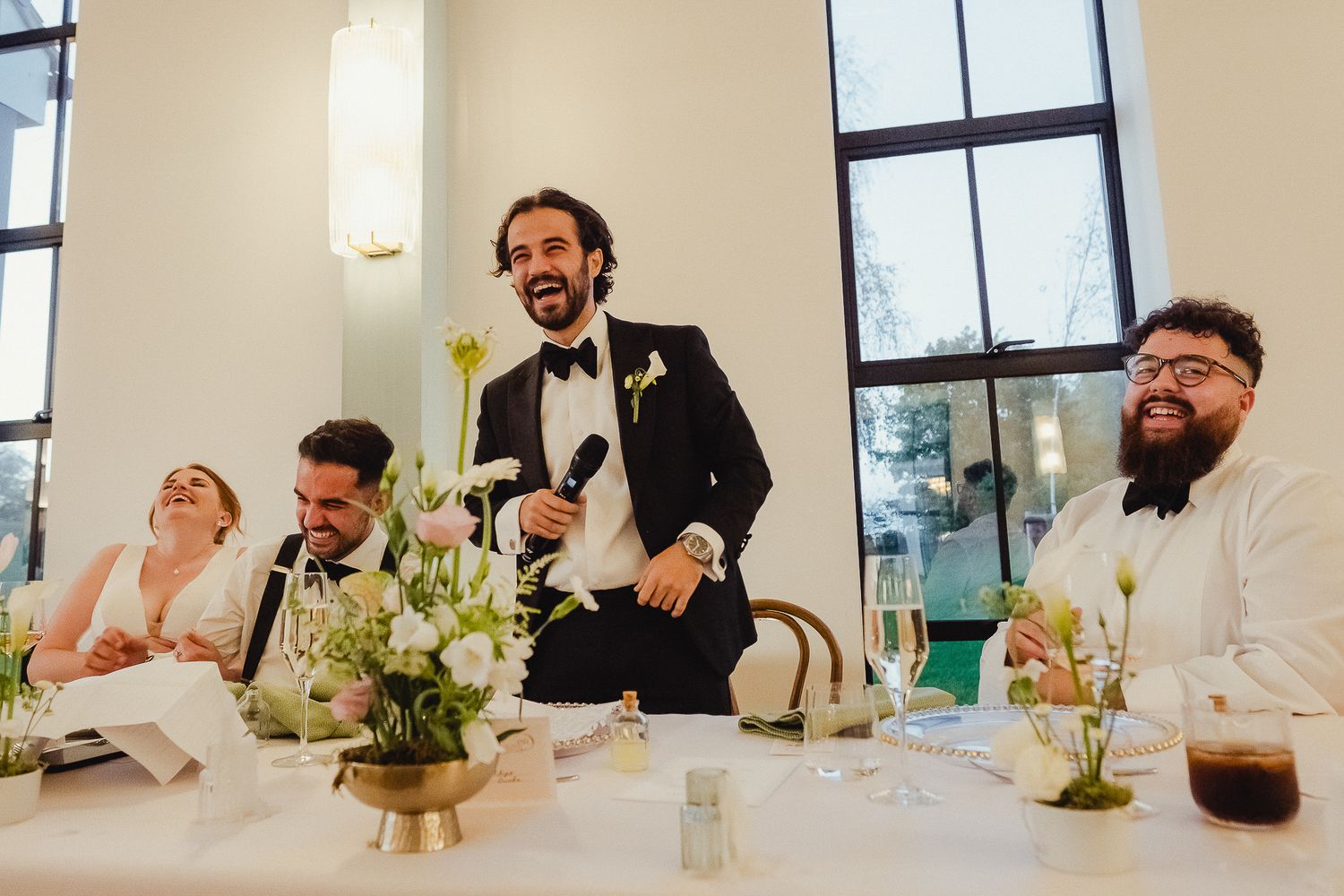Wedding party laughs together at head table during reception with white floral centerpieces and natural light streaming in.