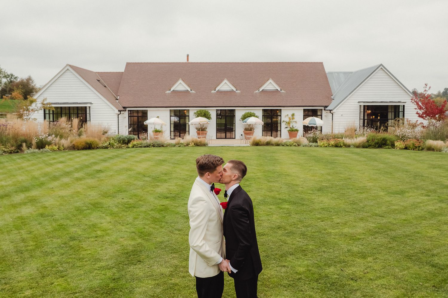 A couple embraces on a grassy lawn with a white building in the background during an autumn day.