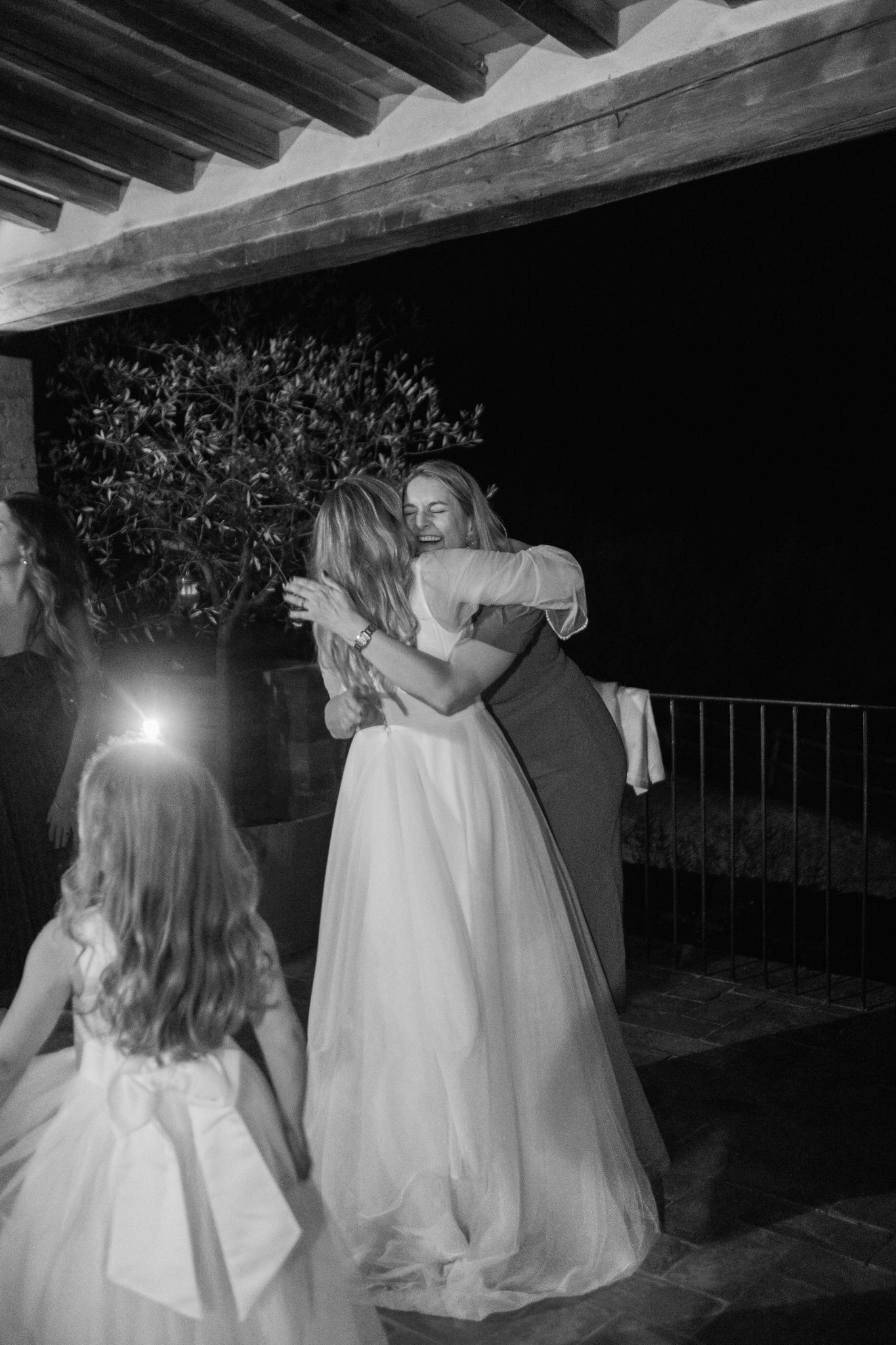 Bride in a flowing white wedding gown embraces a smiling guest on a candlelit terrace while a flower girl looks on.