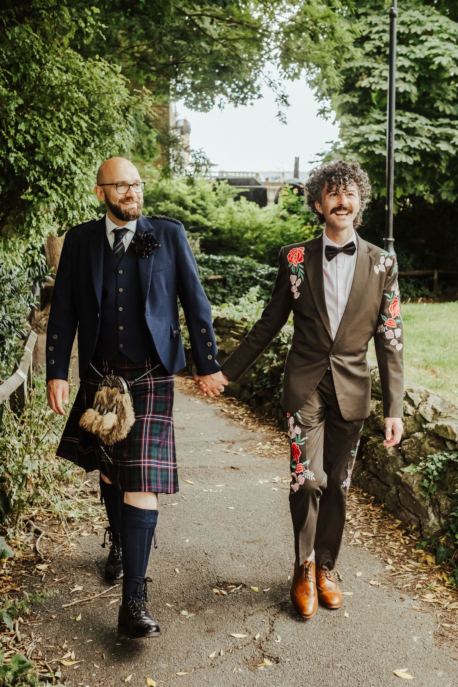 Two grooms holding hands on a leafy garden path, one in a navy jacket and tartan kilt, the other in a floral-embroidered suit and bow tie.