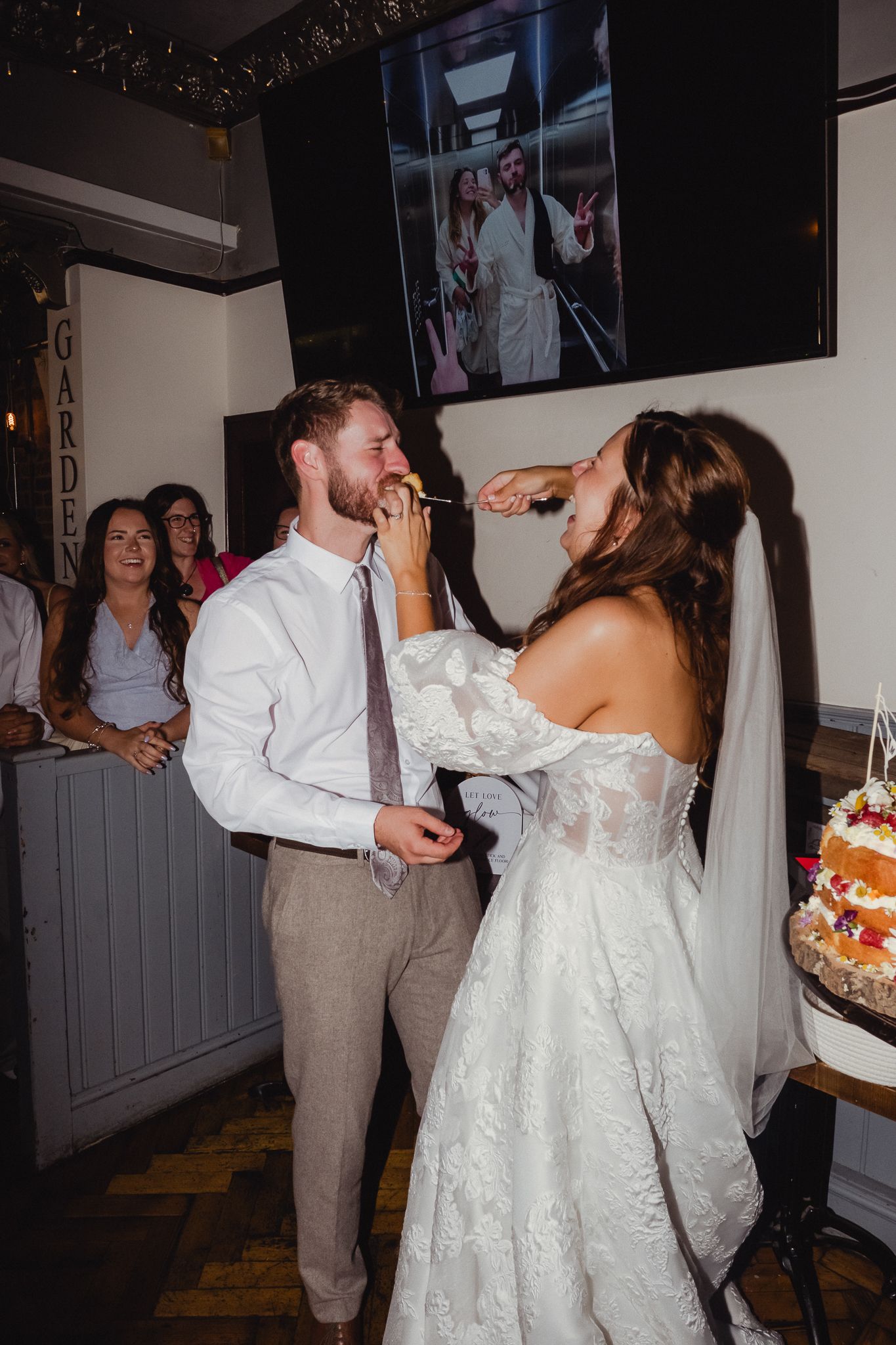 A bride and groom share cake during their wedding reception while guests smile in the background.