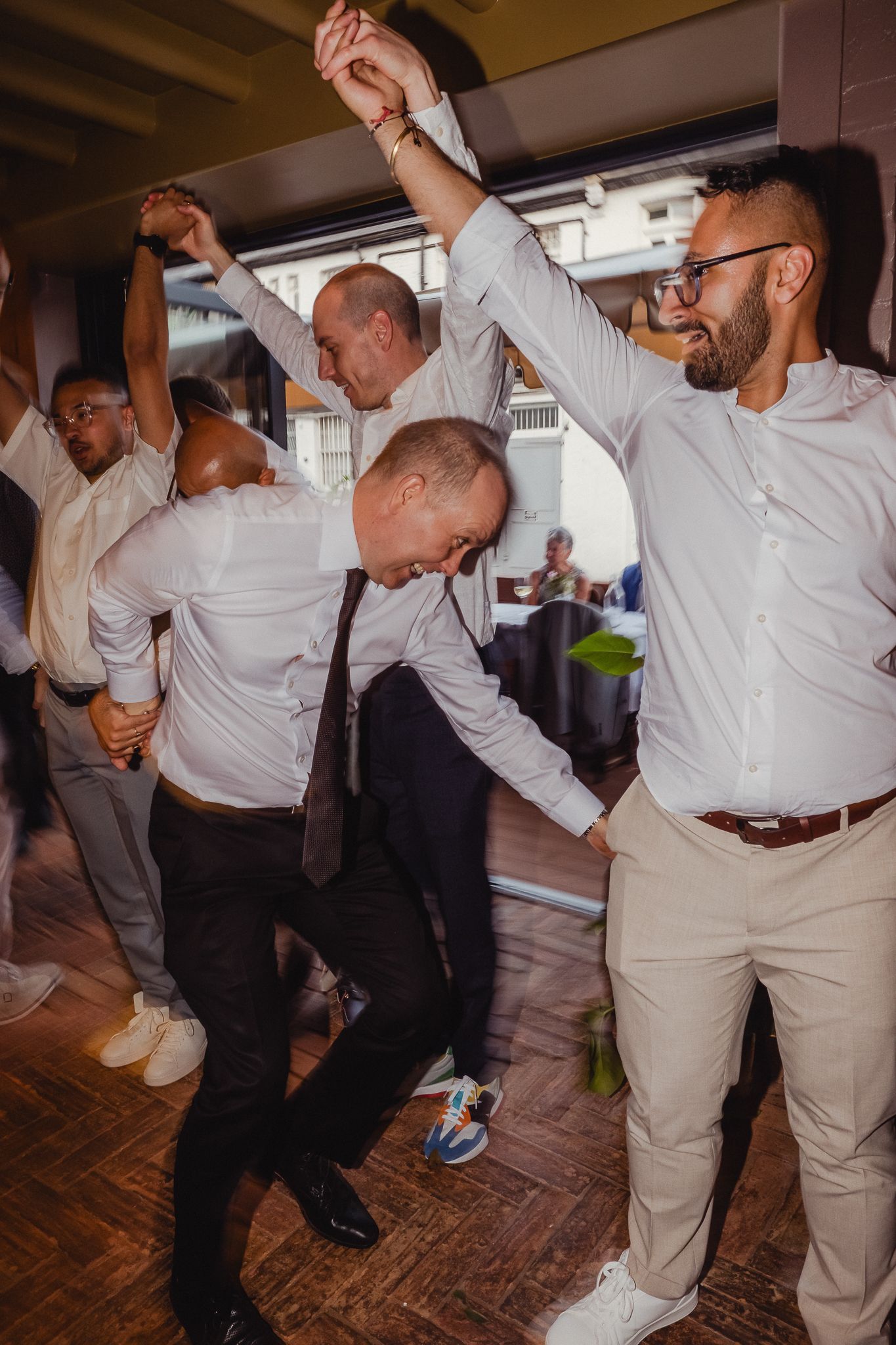 Wedding guests in white shirts and khaki pants dance energetically on a wooden dance floor at a reception celebration.