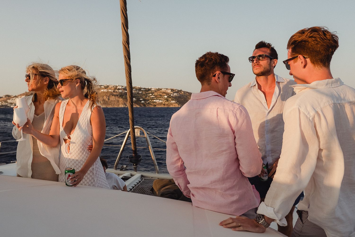 Group of wedding guests enjoying a sunset boat ride along the coastline in elegant white and pink attire.