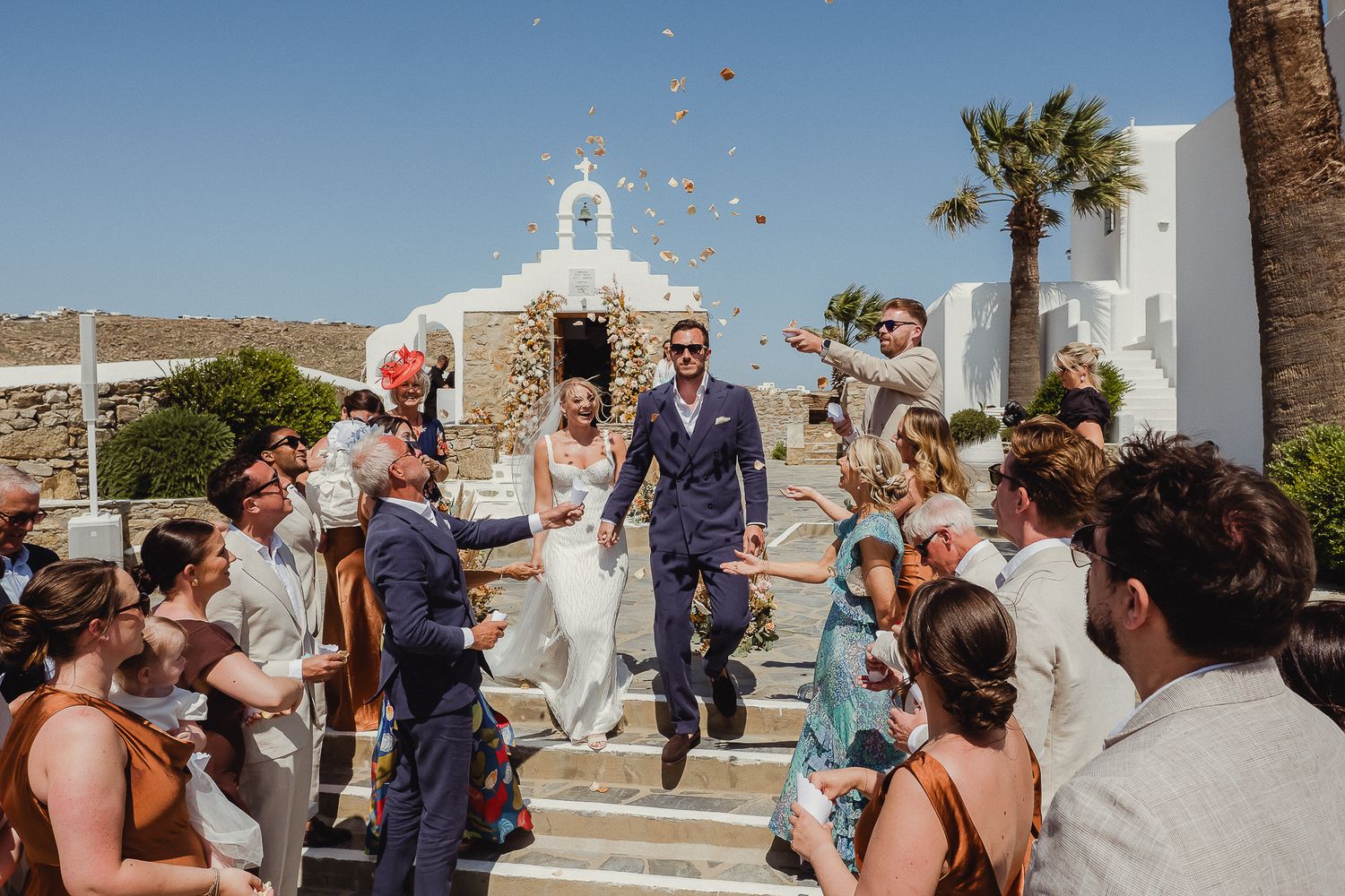 Wedding celebration on white church steps with confetti toss in sunlit Mediterranean setting with palm trees.