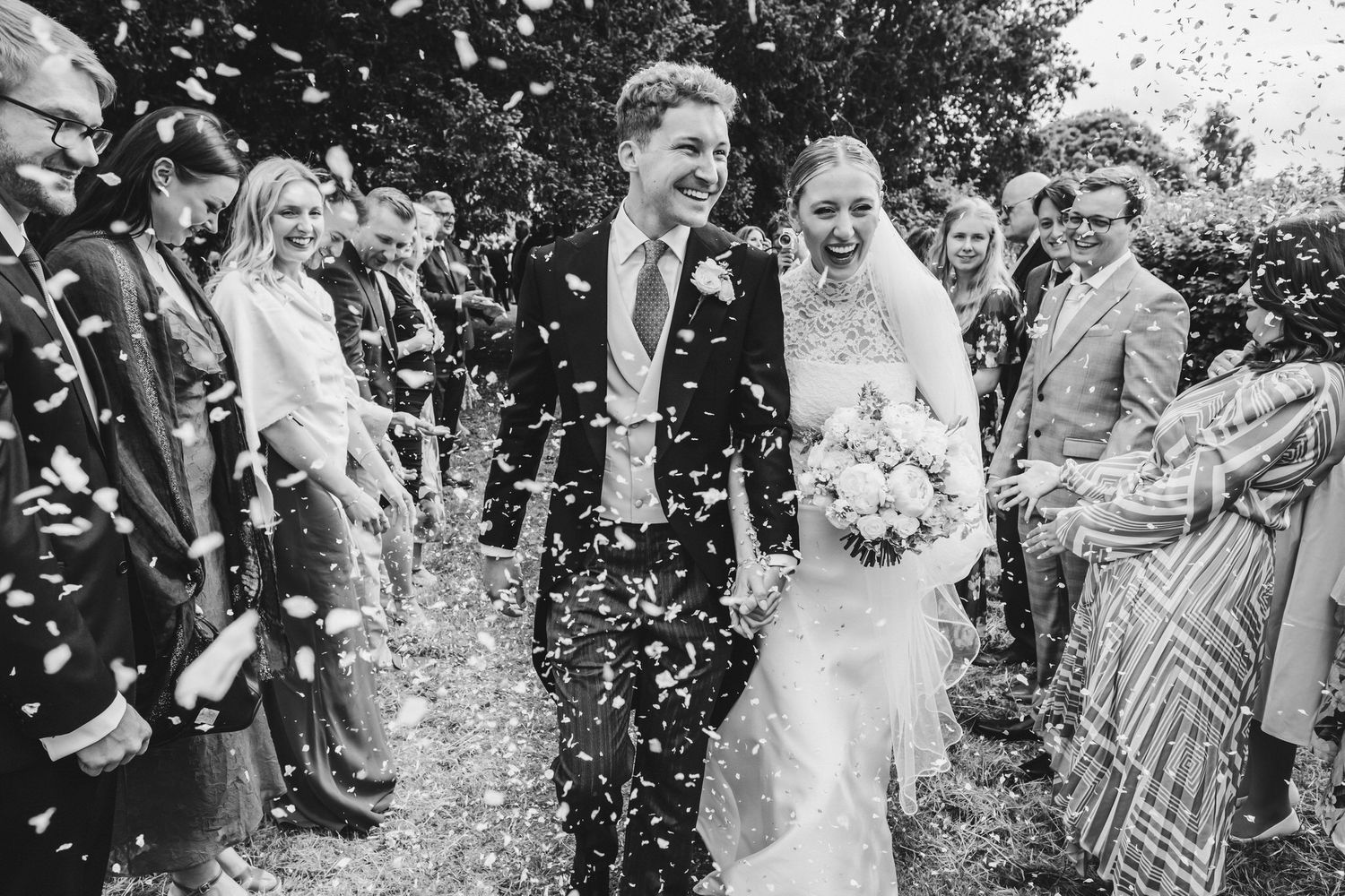 Wedding guests joyfully toss white confetti over a couple during an outdoor celebration at a garden ceremony.