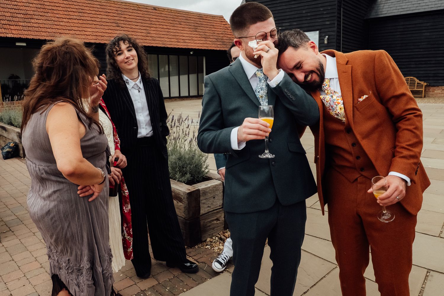 Two men in suits share an emotional moment at a wedding reception while two women look on outside a barn venue.