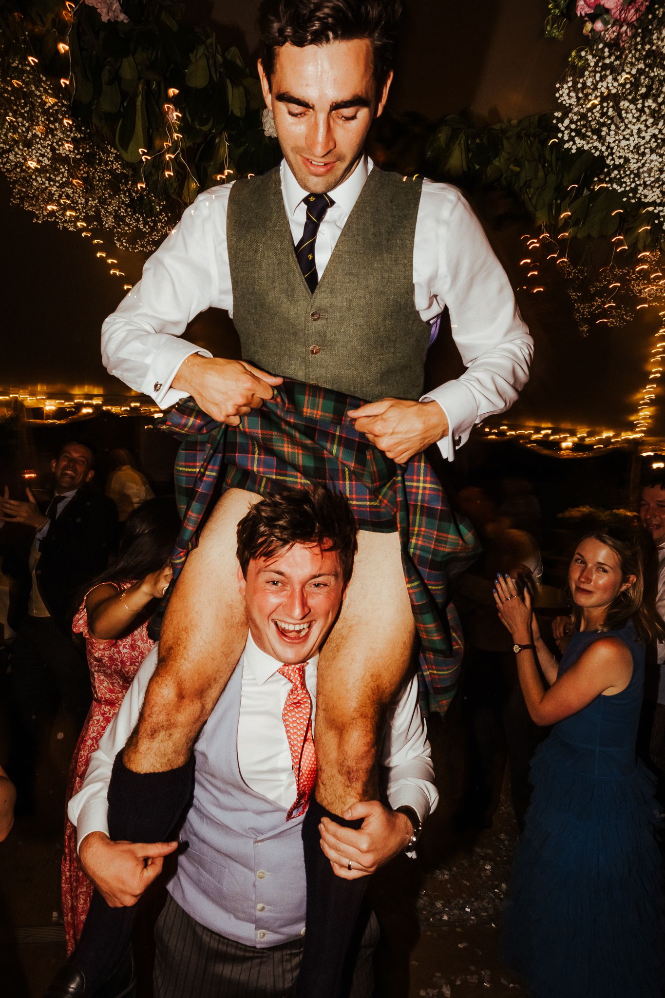 Celebratory moment at a wedding reception with guests lifting someone wearing traditional Scottish attire.