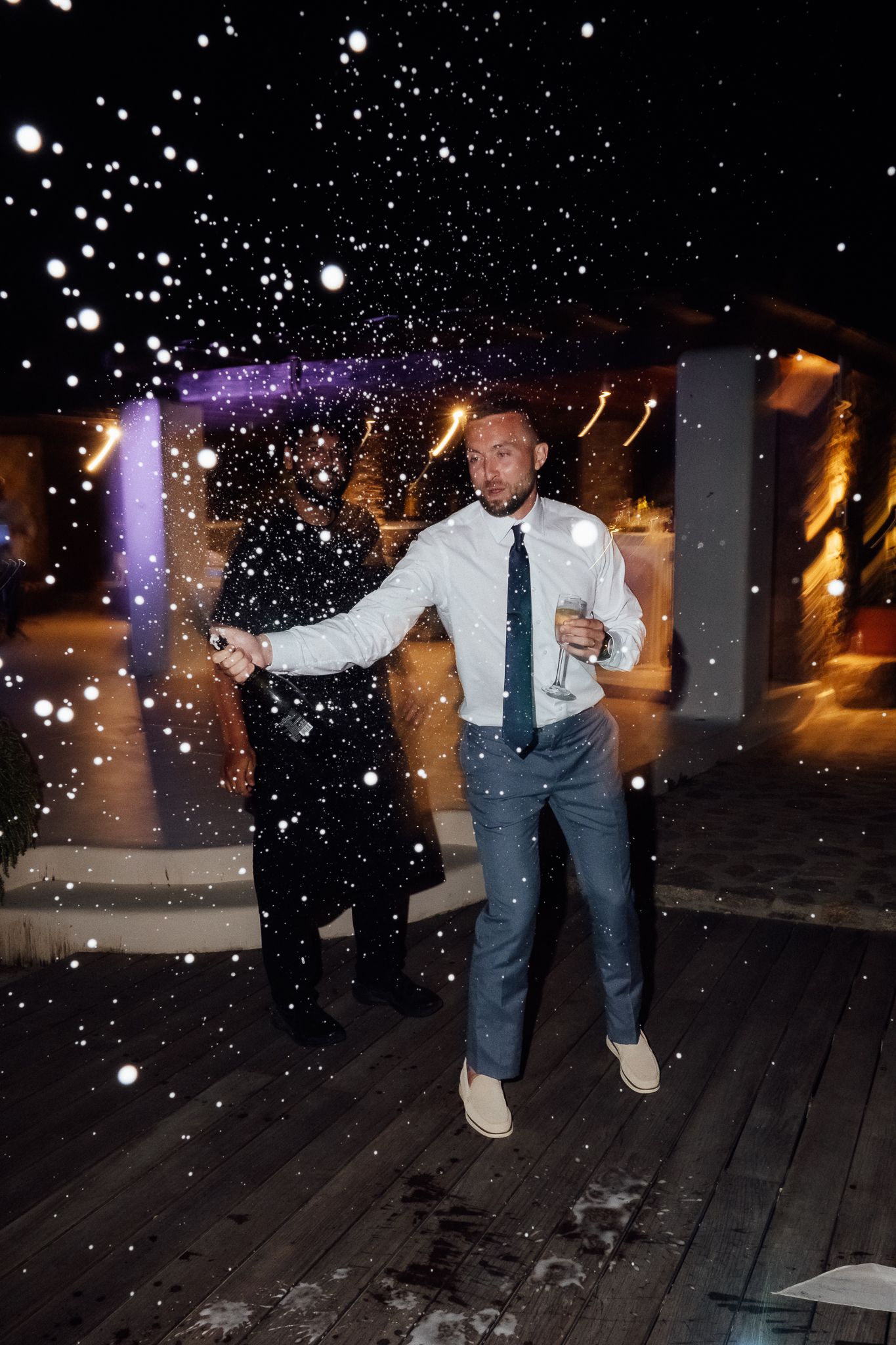 Man in white shirt and tie holding champagne glass outdoors at night with snow falling at a party.