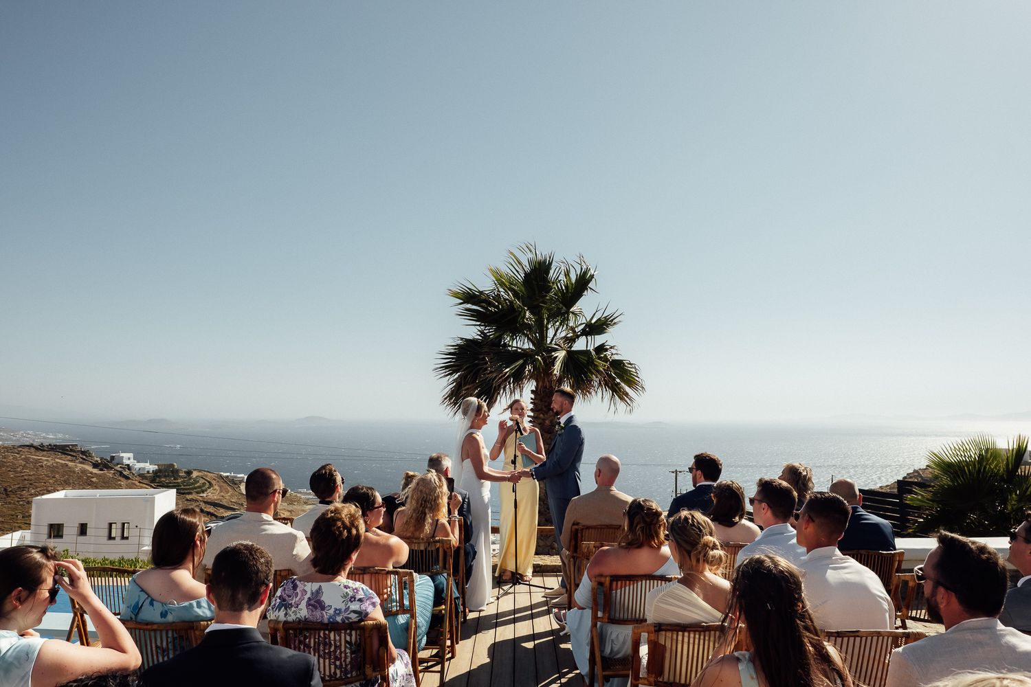 Outdoor wedding ceremony with ocean view, guests seated facing couple at altar near palm tree on sunny day.